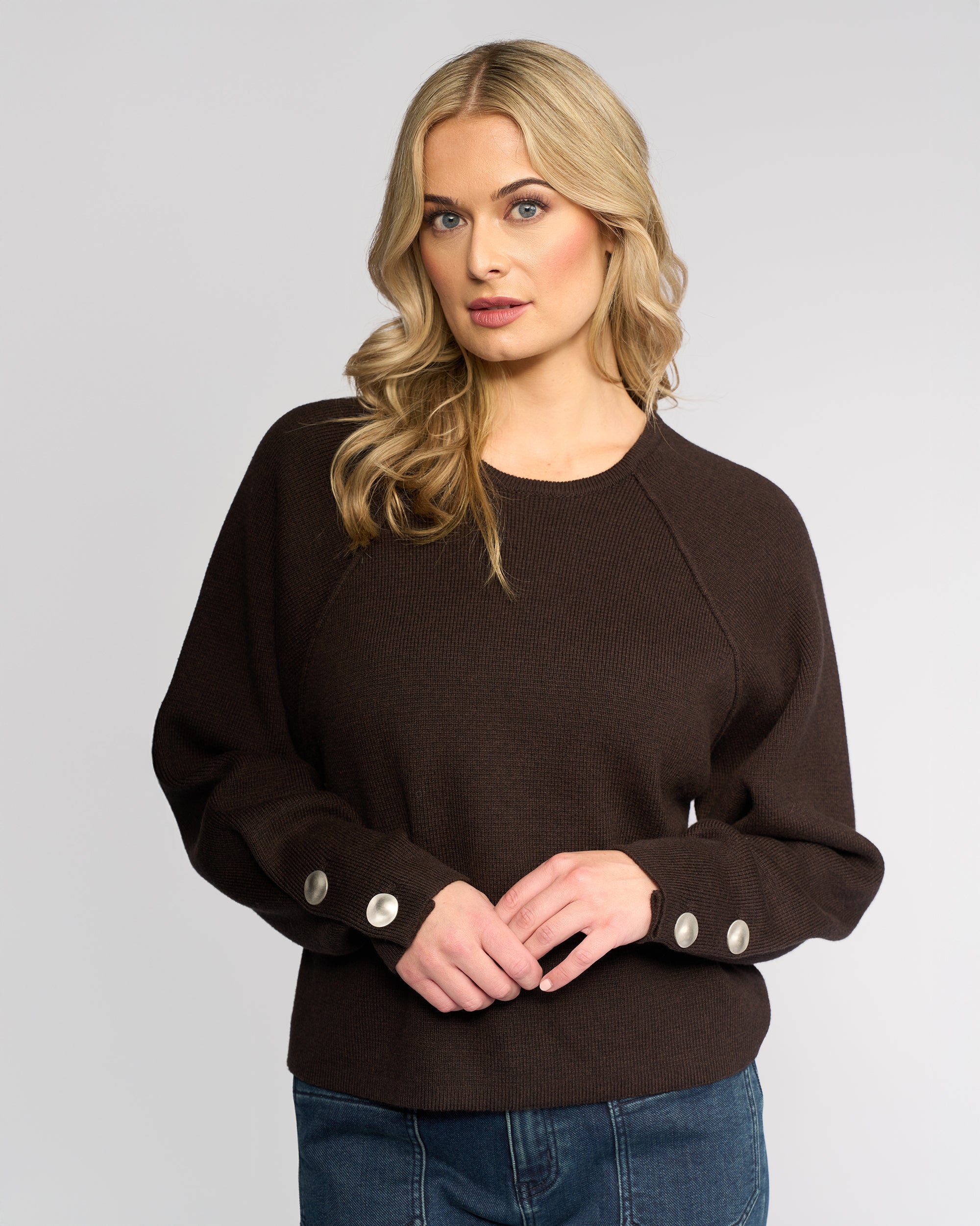 A woman with long blonde hair models the Knewe Label Juno Sweater in chocolate, featuring oversized sleeves and large buttoned cuffs, paired with blue jeans against a light gray background.