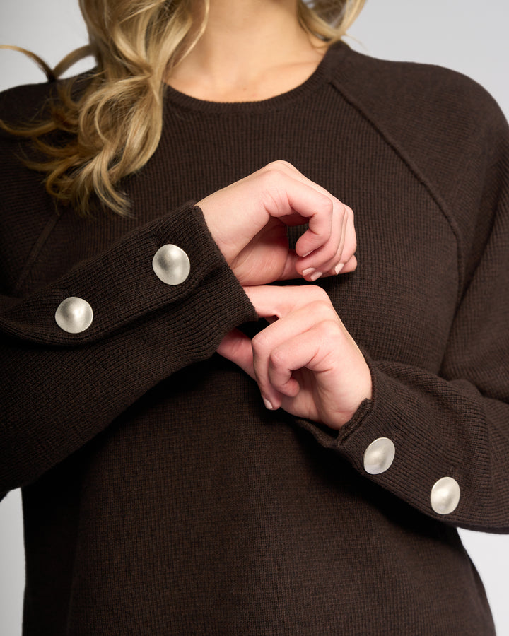 A person wearing Knewe Label’s Juno Sweater in Chocolate—a dark brown ribbed knit with oversized sleeves and silver cuff buttons—is shown from the shoulders down, gently touching one wrist with the other hand.