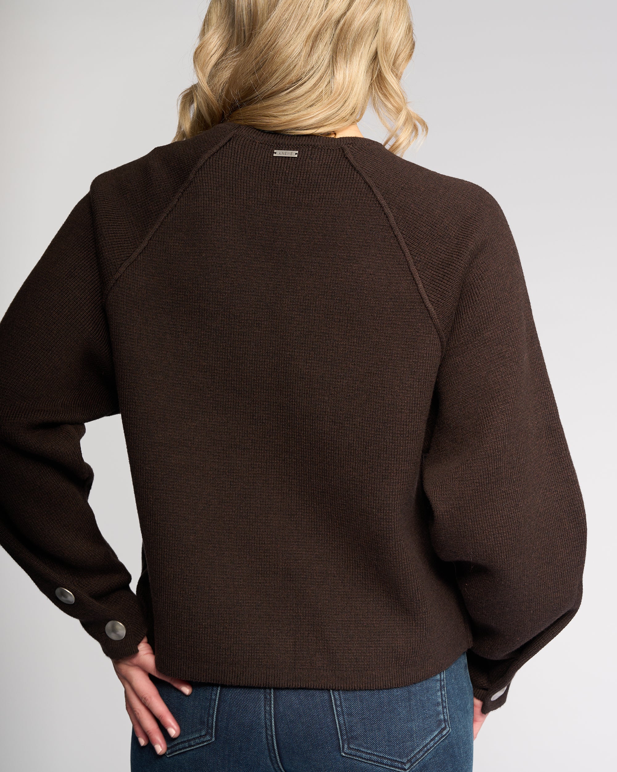 A person with wavy blonde hair is shown from the back wearing the Knewe Label Juno Sweater in Chocolate, featuring oversized sleeves and silver cuff buttons, paired with blue jeans against a plain light background.