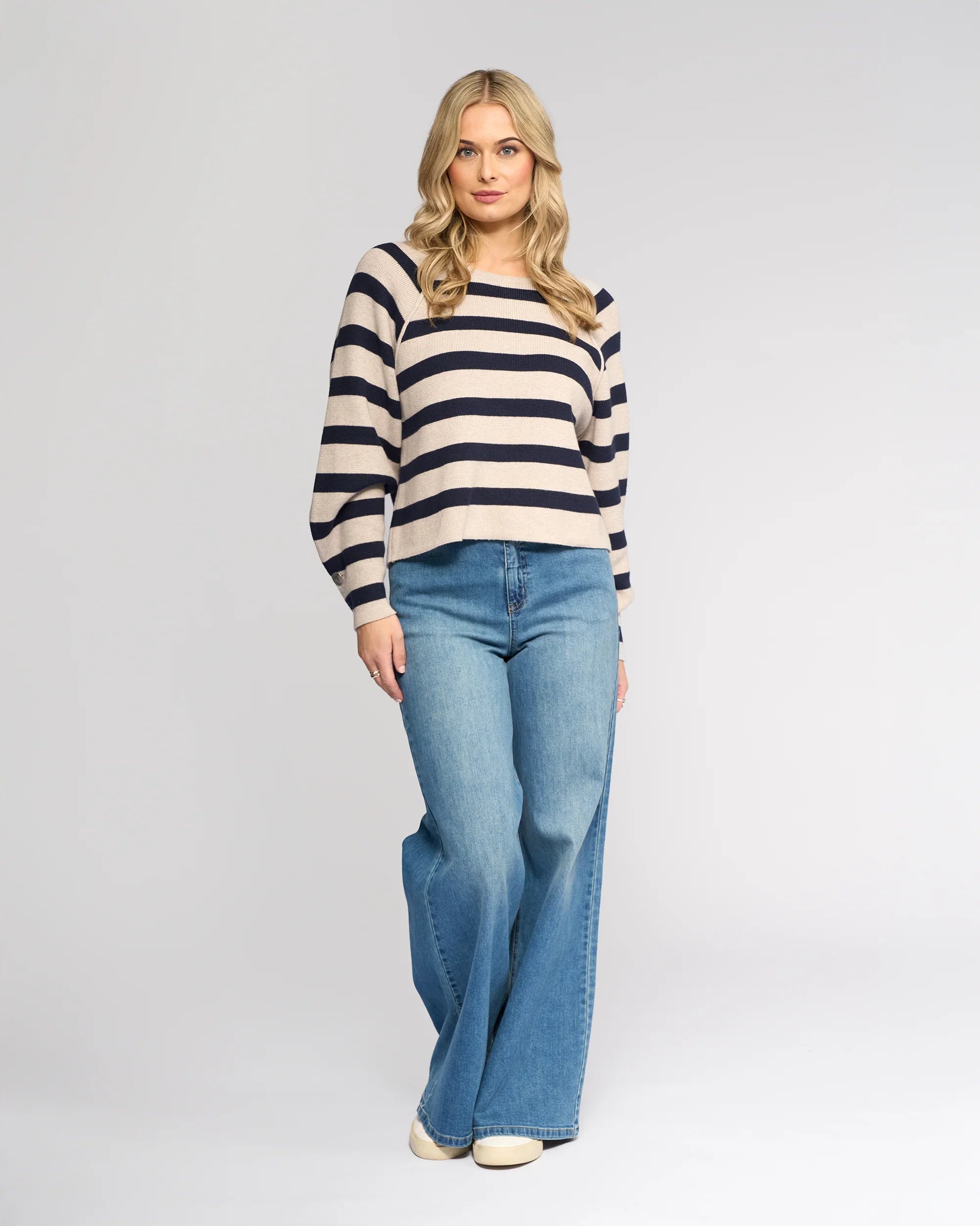 A woman with long blonde hair wears the Knewe Label Juno Sweater in Oat/Ink, featuring cream and navy stripes and oversized sleeves, paired with wide-leg blue jeans. She stands facing forward against a plain light gray background.