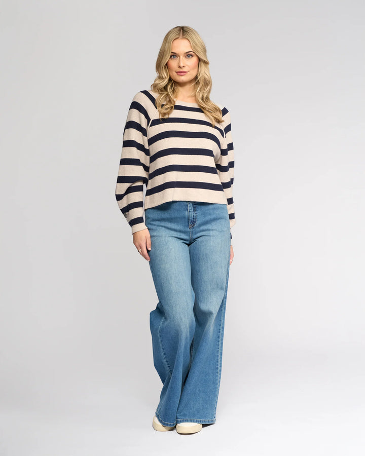 A woman with long blonde hair wears the Knewe Label Juno Sweater in Oat/Ink, featuring cream and navy stripes and oversized sleeves, paired with wide-leg blue jeans. She stands facing forward against a plain light gray background.