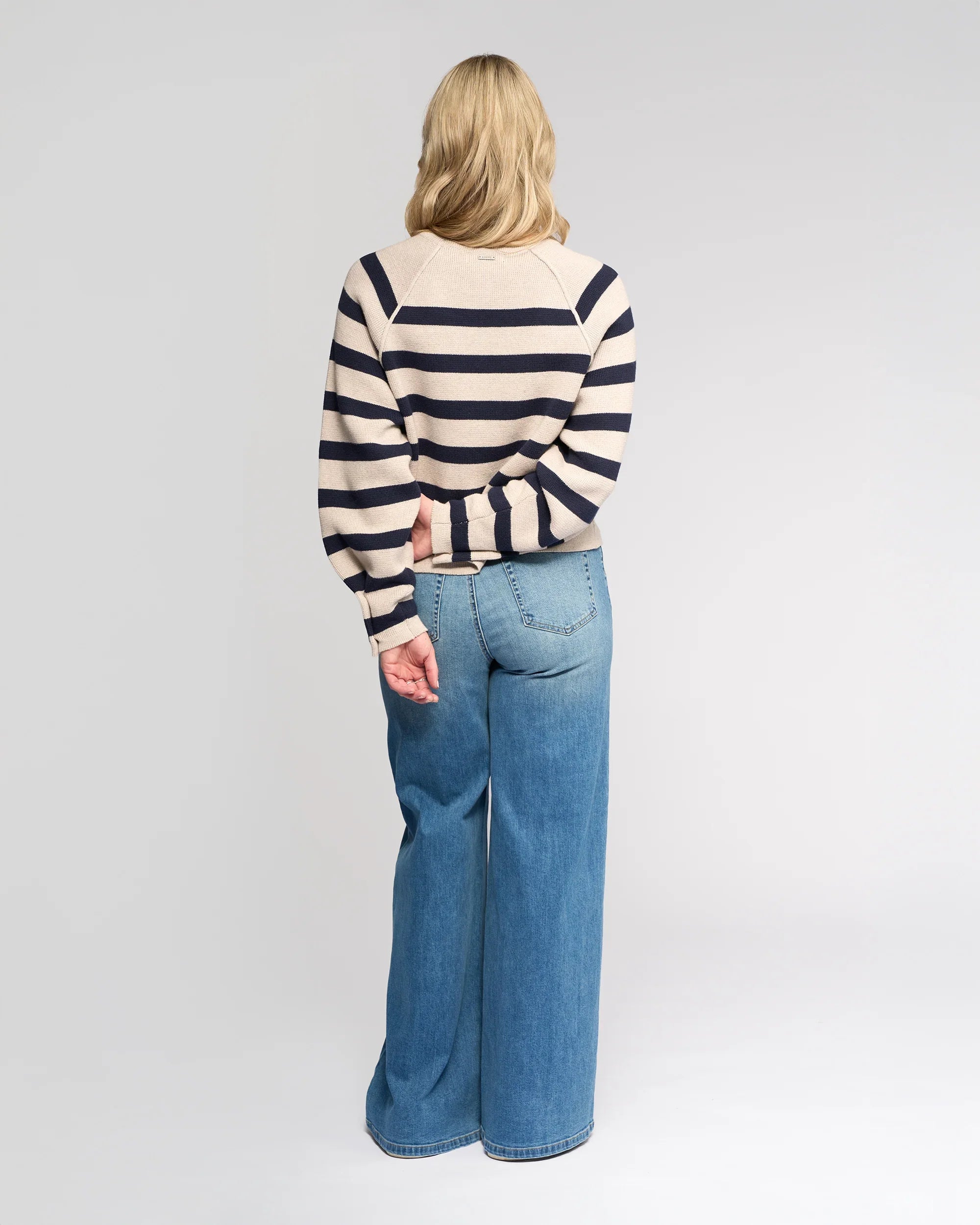 A person with long blonde hair, facing away, wears the Knewe Label Juno Sweater in Oat/Ink with oversized sleeves and high-waisted wide-leg blue jeans. One arm is behind their back. The background is plain and light-colored.
