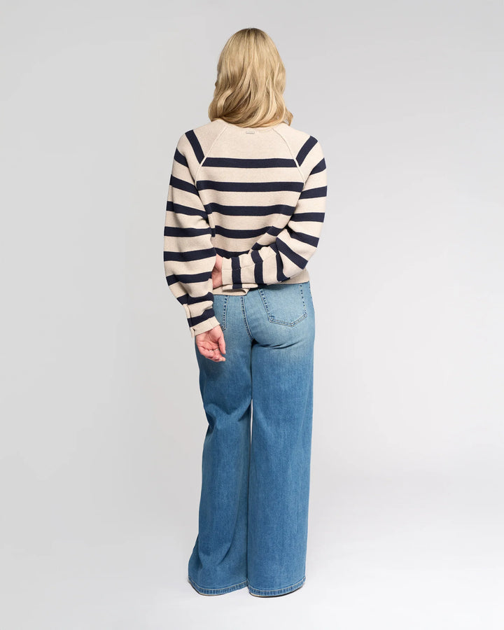 A person with long blonde hair, facing away, wears the Knewe Label Juno Sweater in Oat/Ink with oversized sleeves and high-waisted wide-leg blue jeans. One arm is behind their back. The background is plain and light-colored.