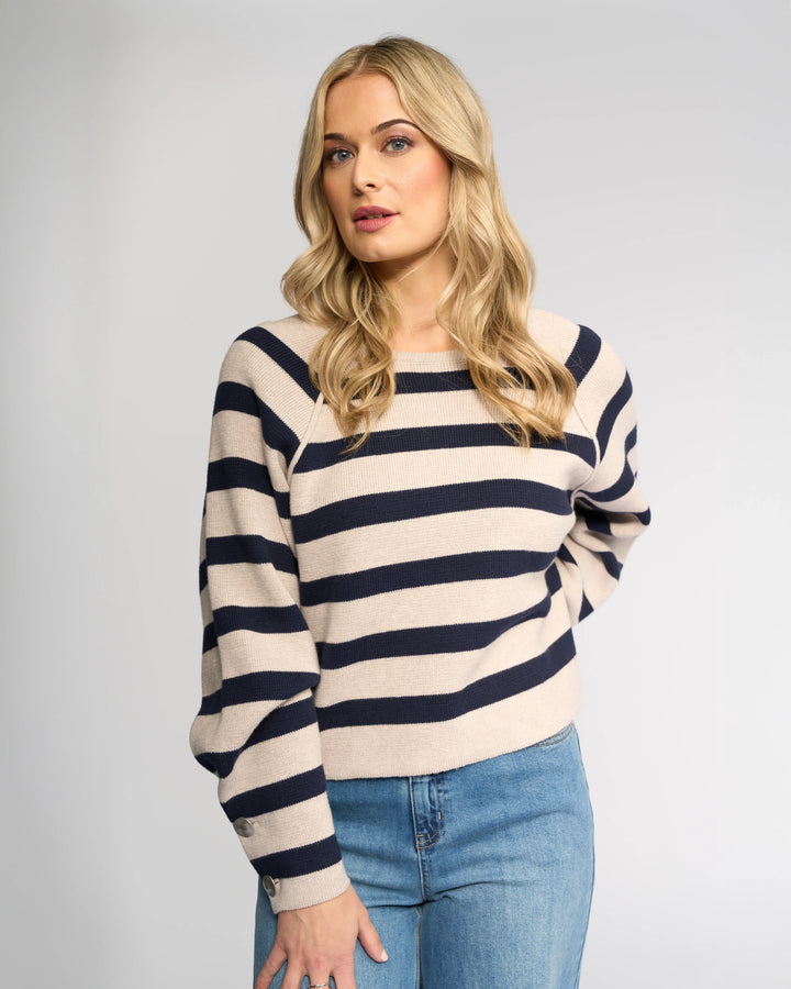 A woman with long blonde hair wears the Knewe Label Juno Sweater in Oat/Ink, featuring navy and beige stripes and oversized sleeves, paired with blue jeans. She stands against a plain light gray background with a neutral expression.