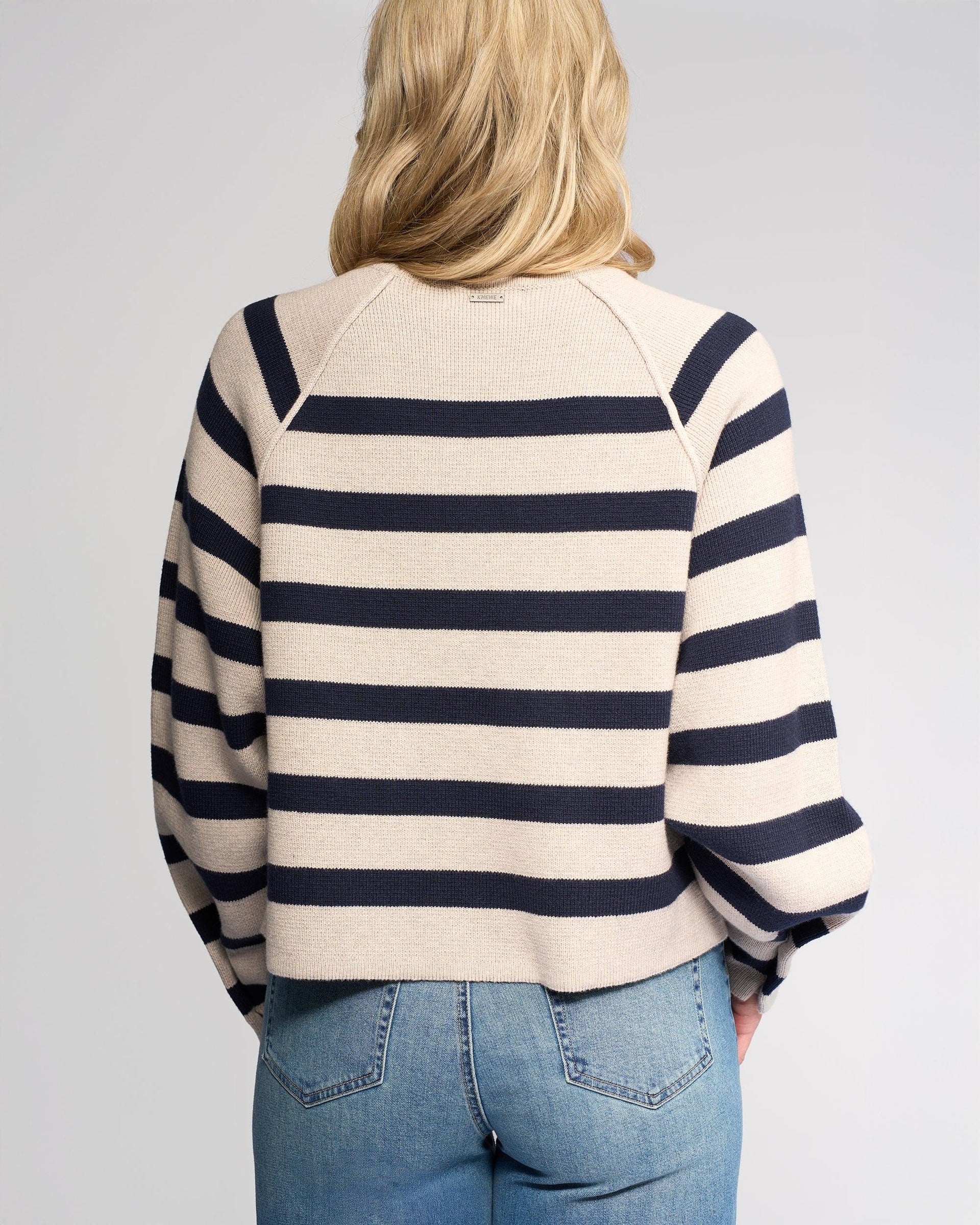 A person with blonde hair, seen from the back, wears the Knewe Label Juno Sweater in Oat/Ink—featuring stripes and oversized sleeves—paired with light blue jeans against a plain light gray background.
