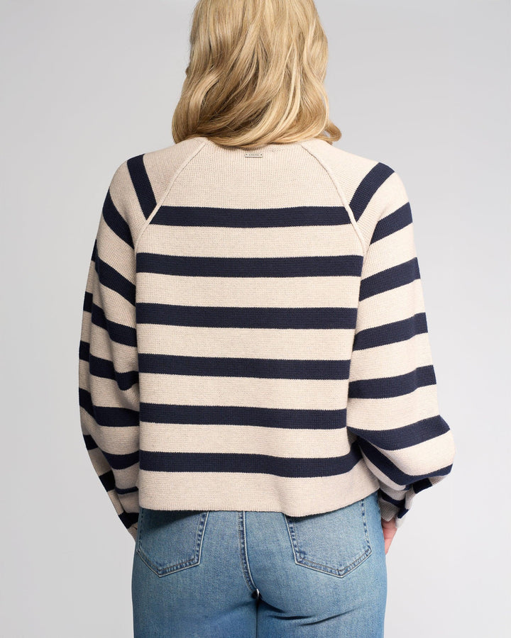 A person with blonde hair, seen from the back, wears the Knewe Label Juno Sweater in Oat/Ink—featuring stripes and oversized sleeves—paired with light blue jeans against a plain light gray background.