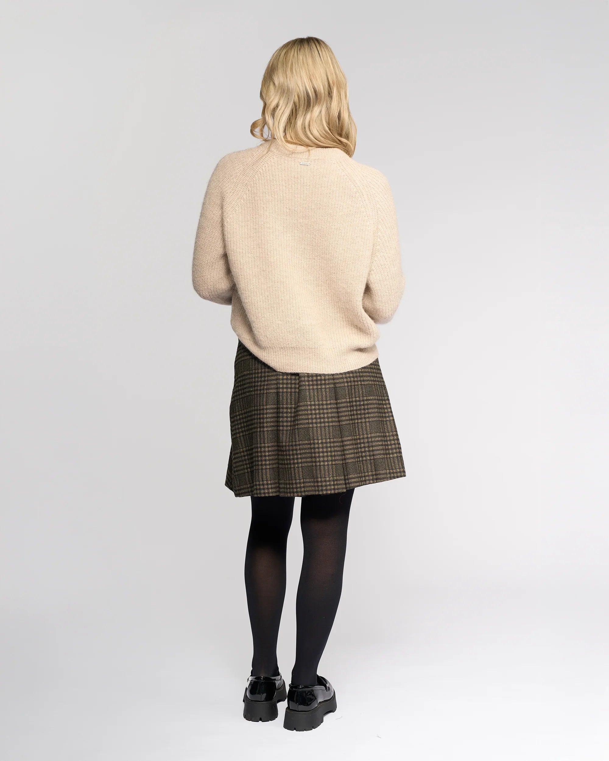 A person with shoulder-length blonde hair faces away, wearing the Knewe Label Fefe Sweater in Oat, paired with a plaid skirt, black tights, and black shoes against a plain light gray background.
