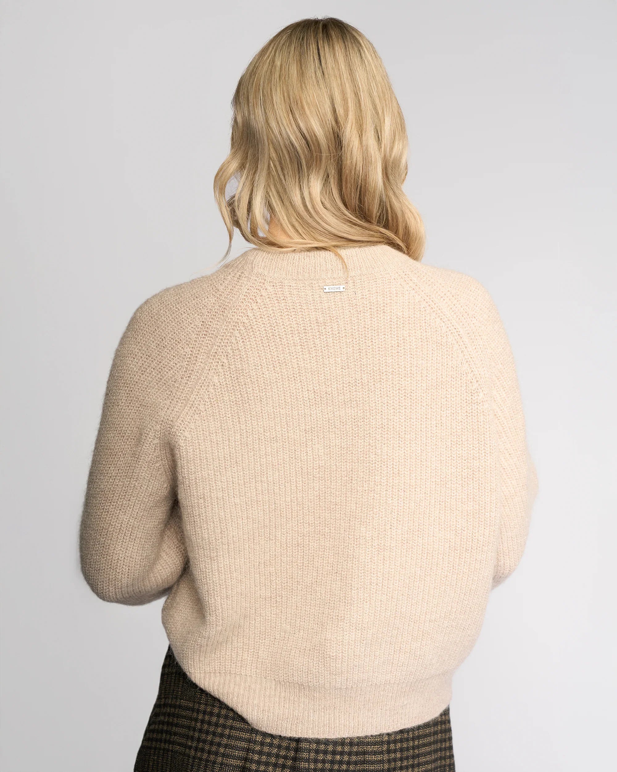 A person with long, wavy blonde hair is seen from the back, wearing the Knewe Label Fefe Sweater in Oat and brown plaid pants against a plain light gray background.