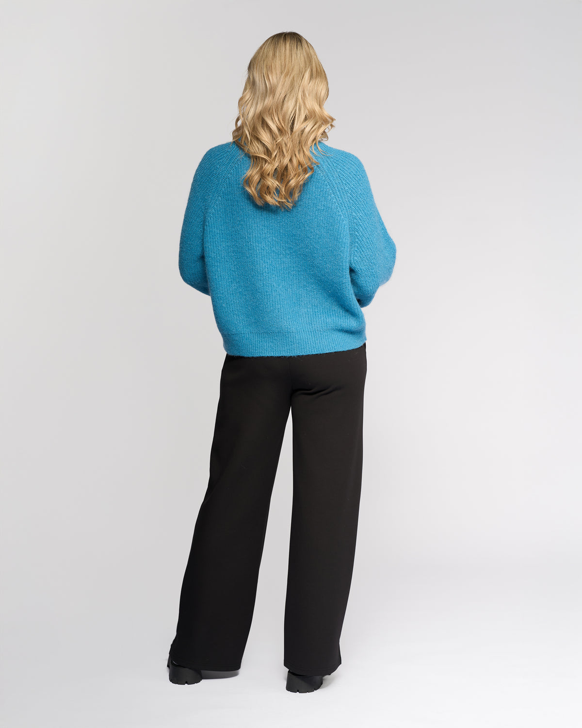 A woman with wavy blonde hair stands facing away, wearing the Knewe Label Fefe Sweater in Ultramarine and black wide-leg pants, against a light gray background.
