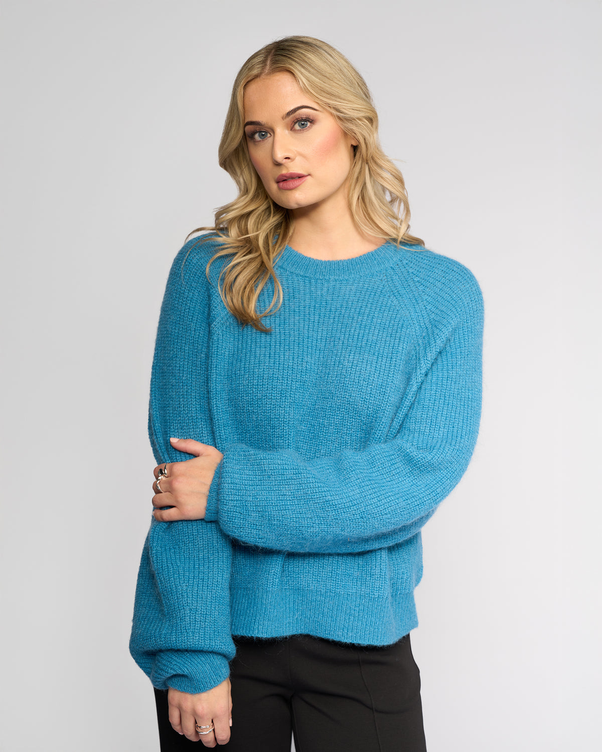 A woman with long blonde hair wears the Knewe Label Fefe Sweater in Ultramarine, paired with black pants, standing against a light gray background and looking to the side with a relaxed expression.