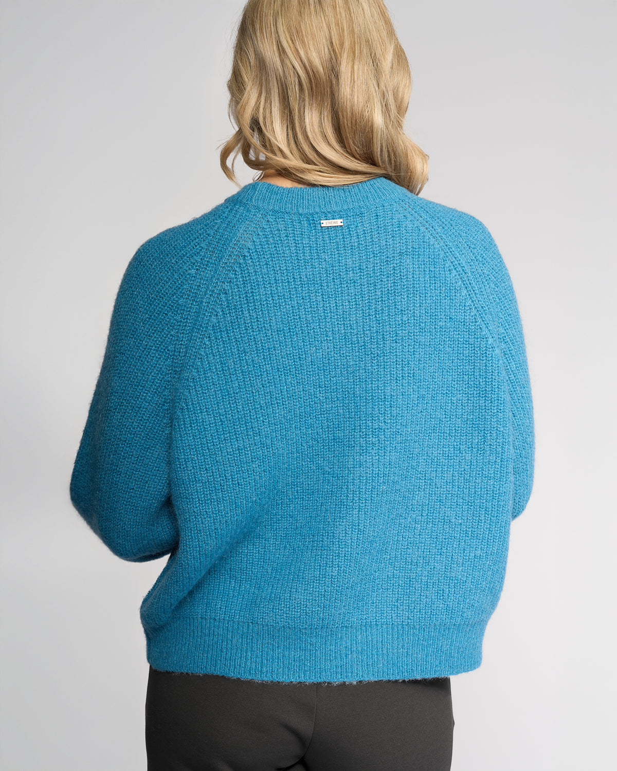 A person with shoulder-length blonde hair is seen from behind wearing the Knewe Label Fefe Sweater in Ultramarine paired with black pants against a plain light background.