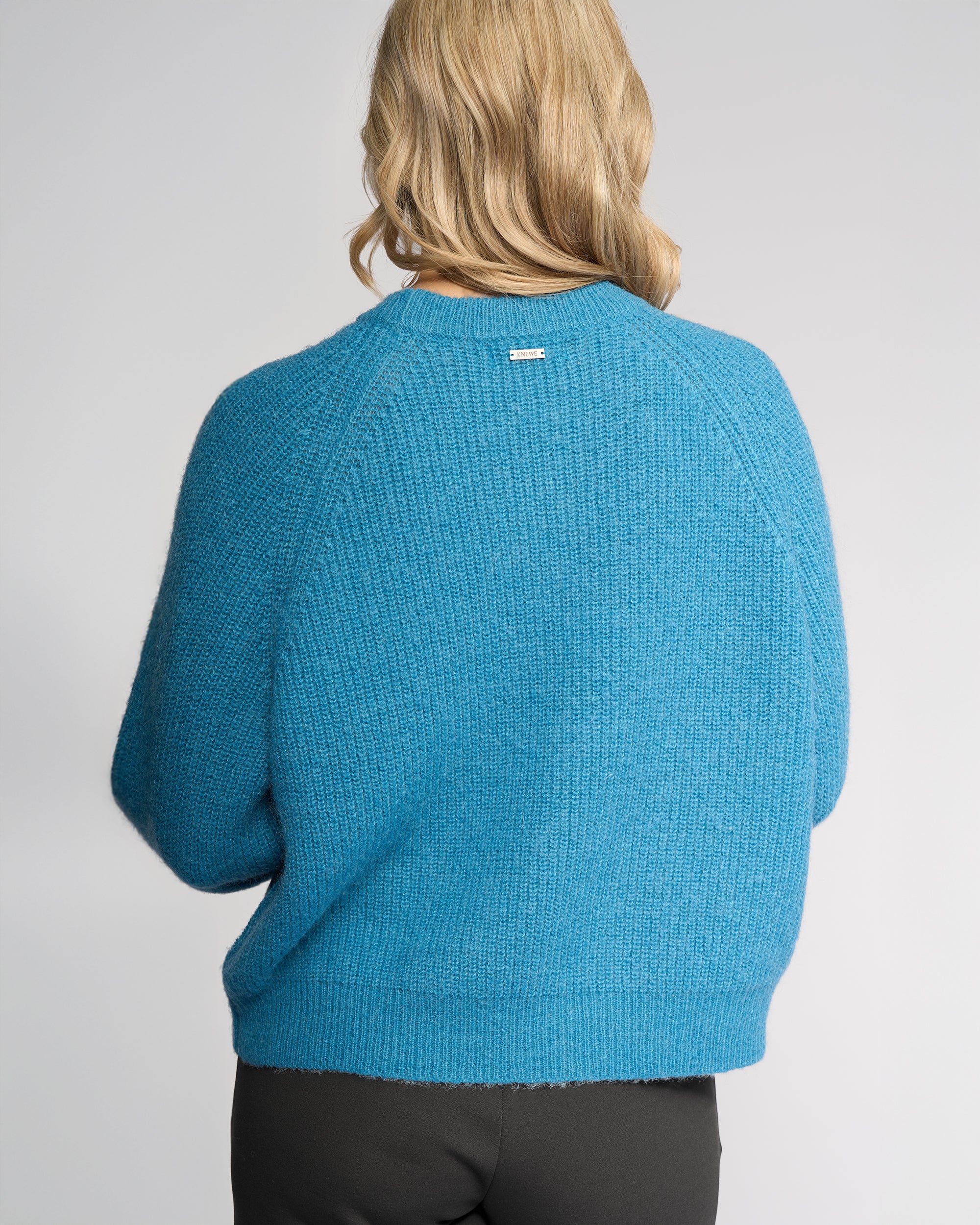 A person with shoulder-length blonde hair is seen from behind wearing the Knewe Label Fefe Sweater in Ultramarine paired with black pants against a plain light background.