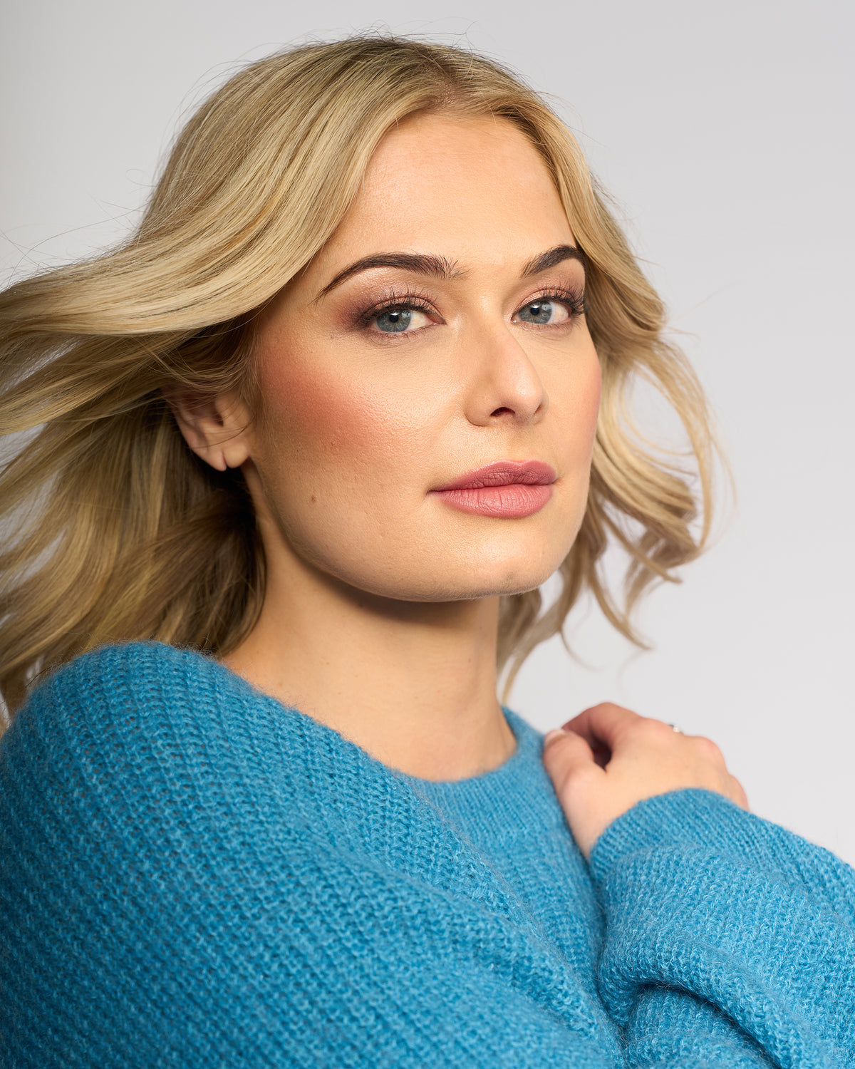 A woman with light blonde hair and natural makeup models the Knewe Label Fefe Sweater in Ultramarine, confidently posing against a plain light background.