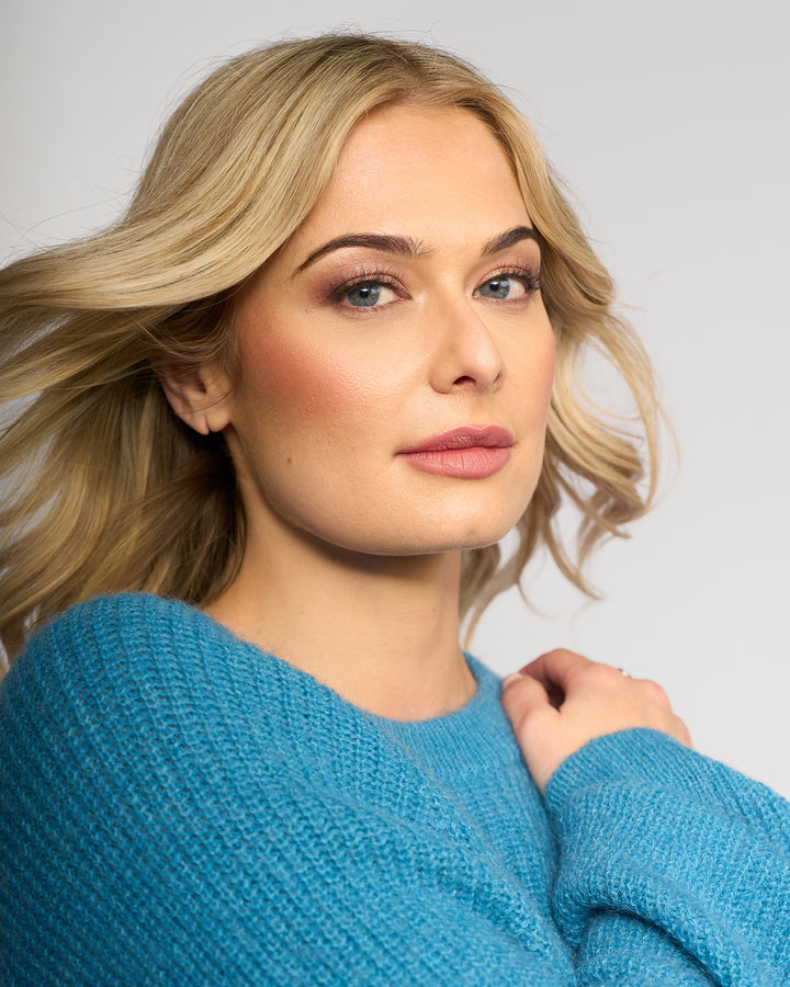 A woman with light blonde hair and natural makeup models the Knewe Label Fefe Sweater in Ultramarine, confidently posing against a plain light background.