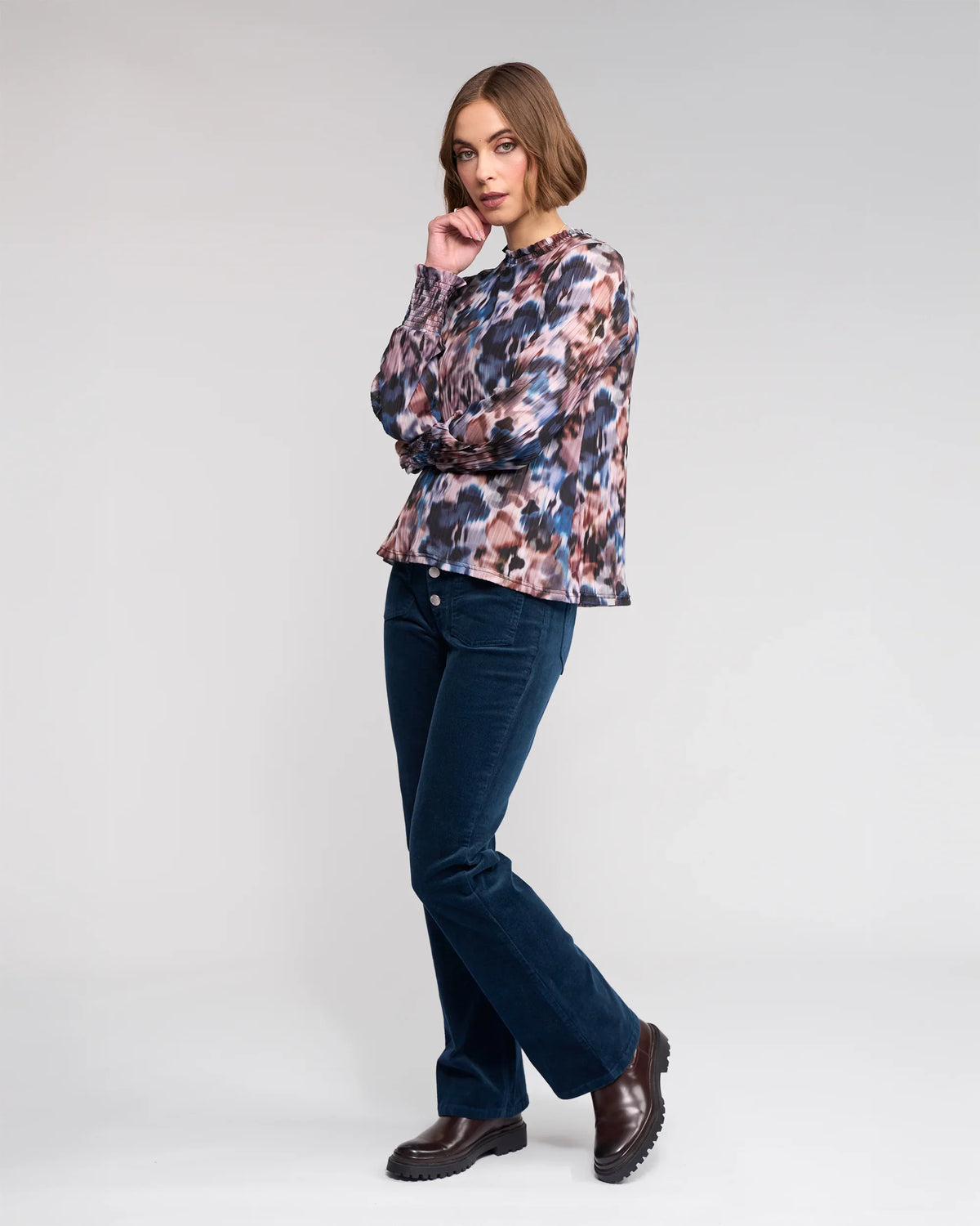A woman with short brown hair poses against a plain background, wearing the Knewe Label Zimi Top - Celine, dark blue corduroy pants, and dark brown boots. She is looking at the camera.
