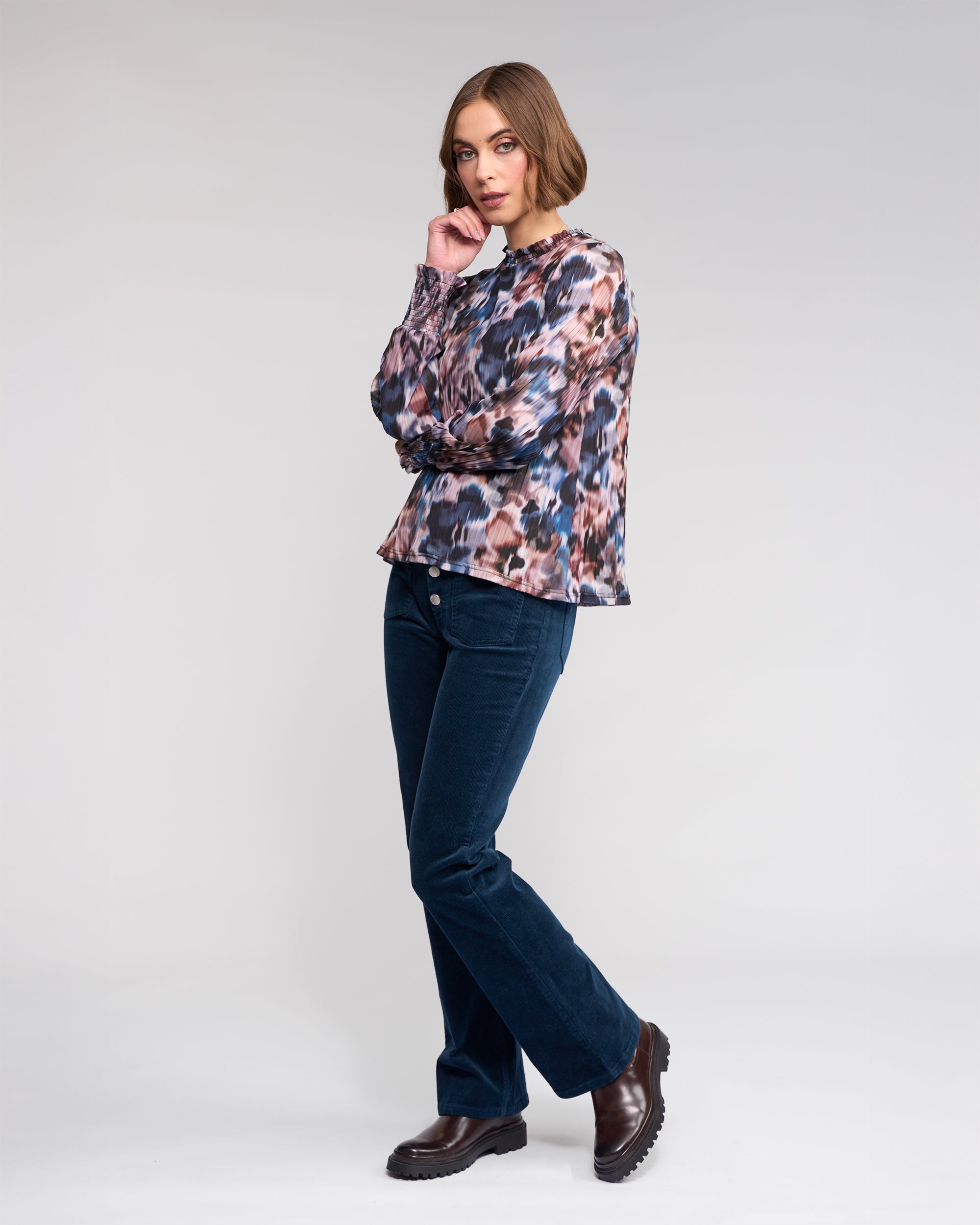 A woman with short brown hair poses against a plain background, wearing the Knewe Label Zimi Top - Celine, dark blue corduroy pants, and dark brown boots. She is looking at the camera.