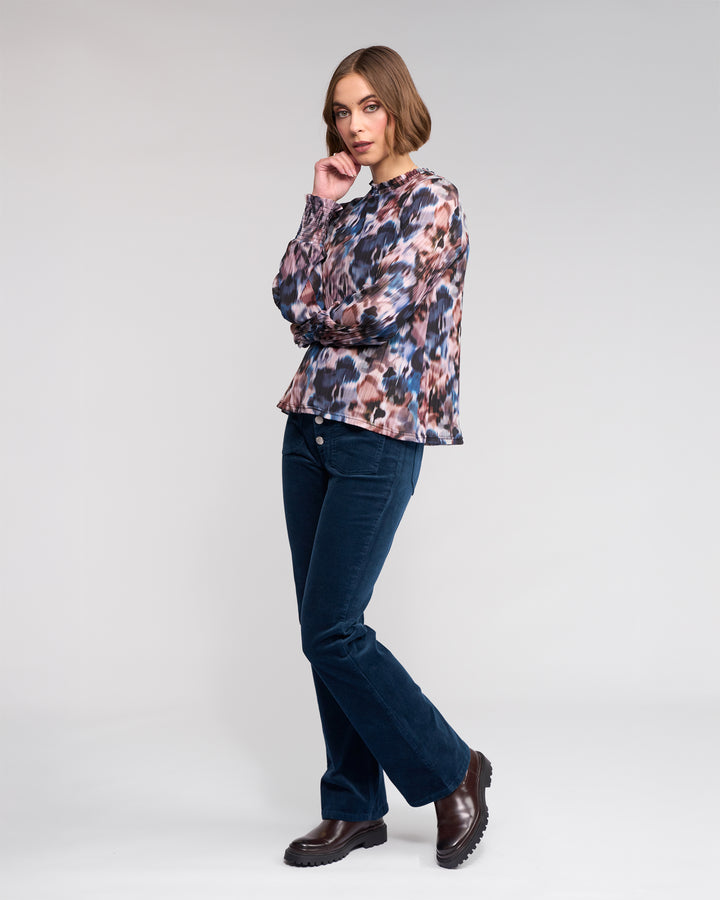 A woman with short brown hair poses against a plain background, wearing the Knewe Label Zimi Top - Celine, dark blue corduroy pants, and dark brown boots. She is looking at the camera.