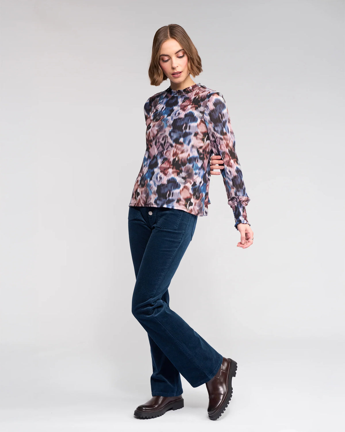 A person with chin-length brown hair wears the Knewe Label Zimi Top - Celine, paired with dark blue corduroy pants and brown boots, standing on one leg with the other slightly bent against a plain light background.