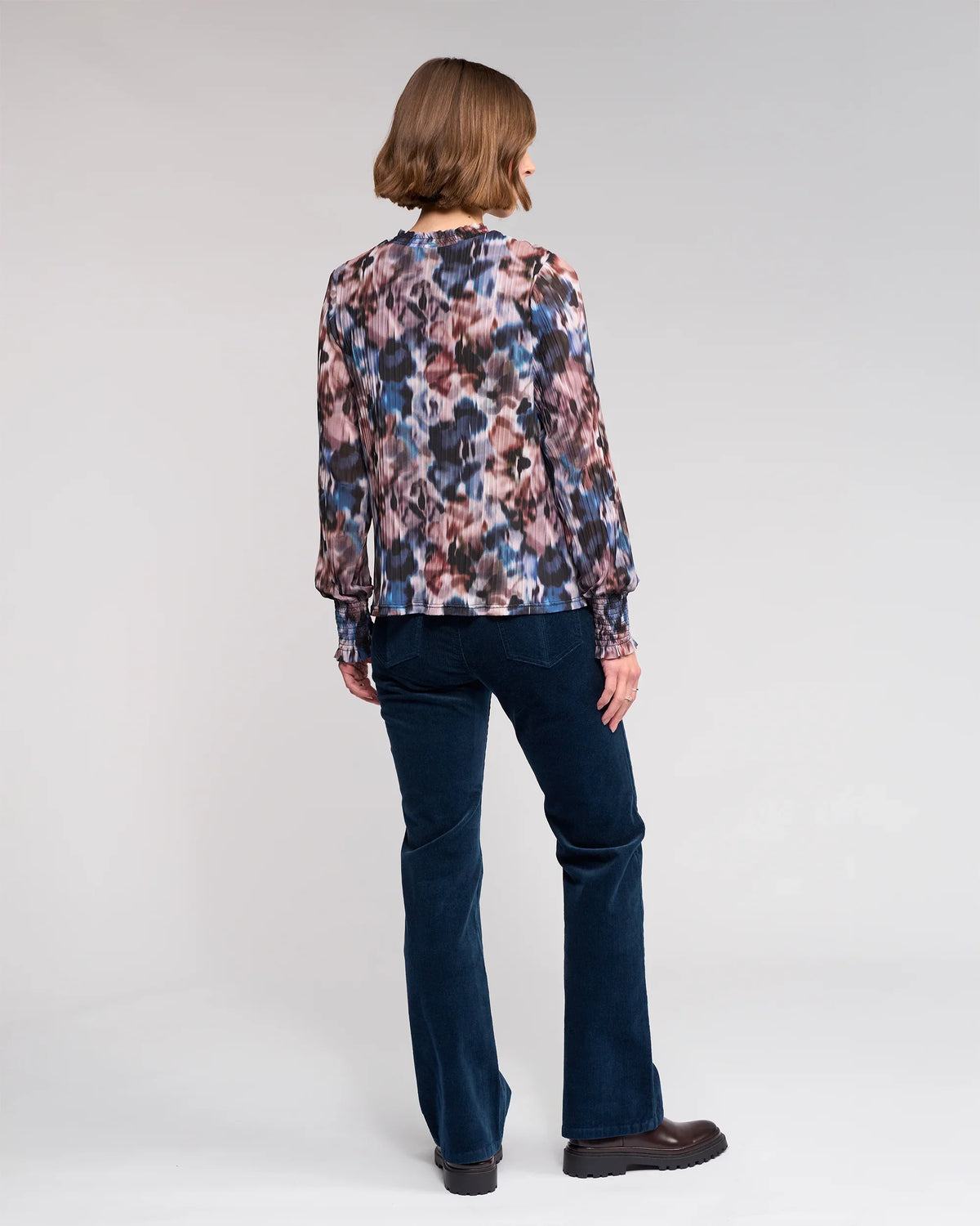 A person with a chin-length bob stands facing away against a light gray background, wearing the Knewe Label Zimi Top - Celine paired with dark blue pants and dark shoes.