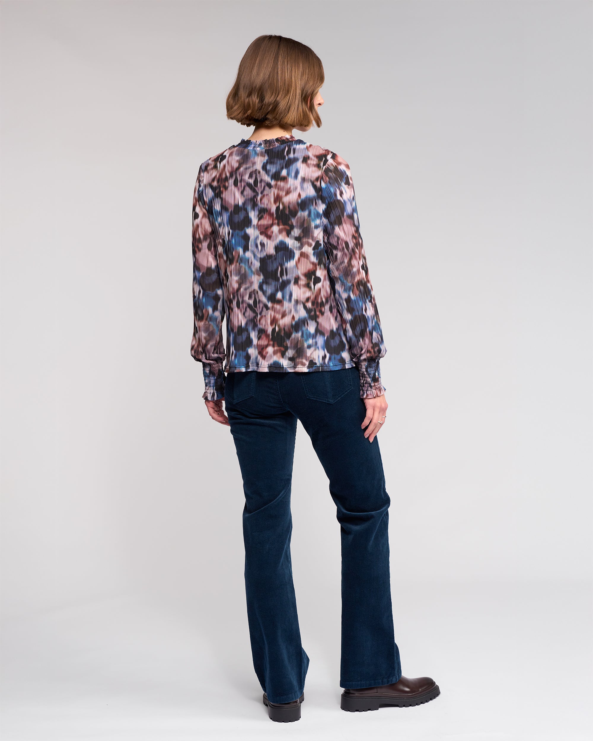 A person with a chin-length bob stands facing away against a light gray background, wearing the Knewe Label Zimi Top - Celine paired with dark blue pants and dark shoes.