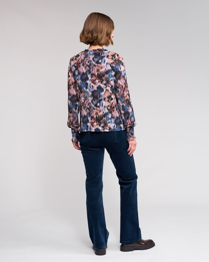 A person with a chin-length bob stands facing away against a light gray background, wearing the Knewe Label Zimi Top - Celine paired with dark blue pants and dark shoes.