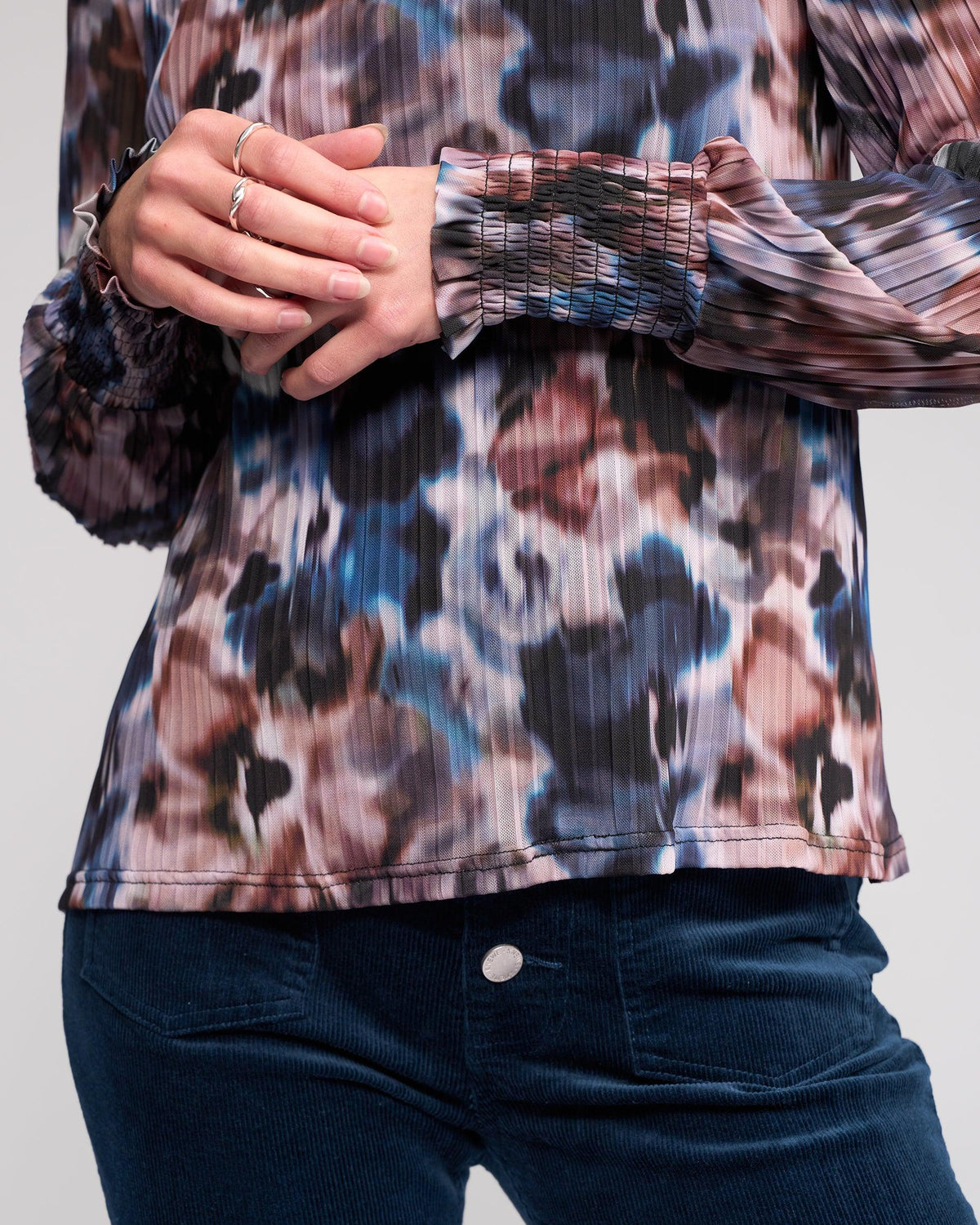 A person wearing the Knewe Label Zimi Top - Celine, a long sleeve pleated blouse with an abstract print, paired with blue corduroy pants and their hands gently clasped at the waist.