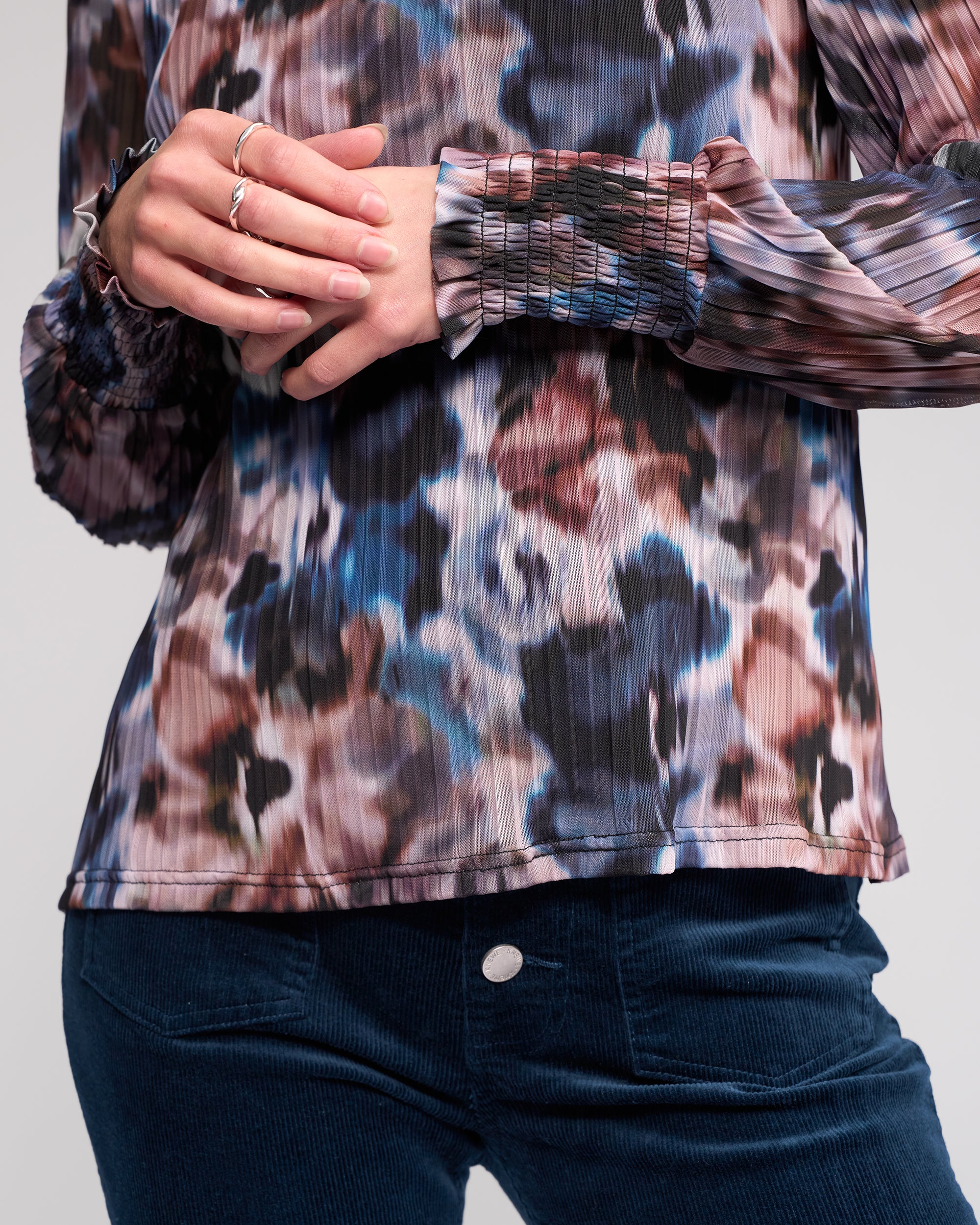A person wearing the Knewe Label Zimi Top - Celine, a long sleeve pleated blouse with an abstract print, paired with blue corduroy pants and their hands gently clasped at the waist.