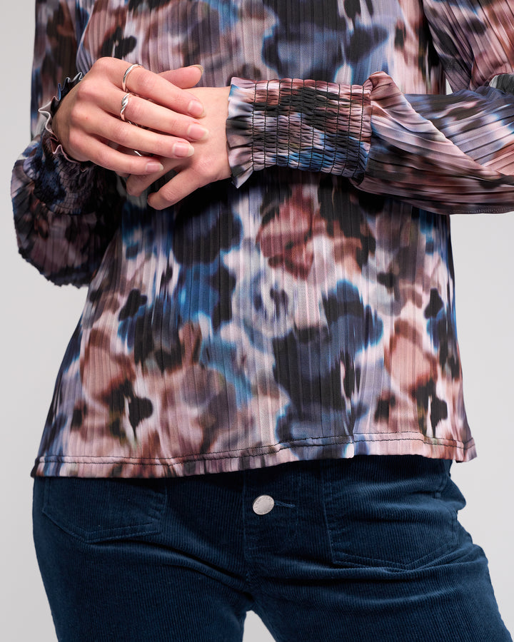 A person wearing the Knewe Label Zimi Top - Celine, a long sleeve pleated blouse with an abstract print, paired with blue corduroy pants and their hands gently clasped at the waist.