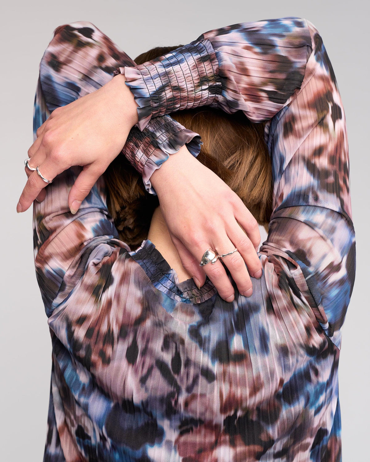 A person faces away, arms raised and crossed behind their head, showcasing silver rings while wearing the Knewe Label Zimi Top - Celine, a long-sleeved blouse with an abstract print.
