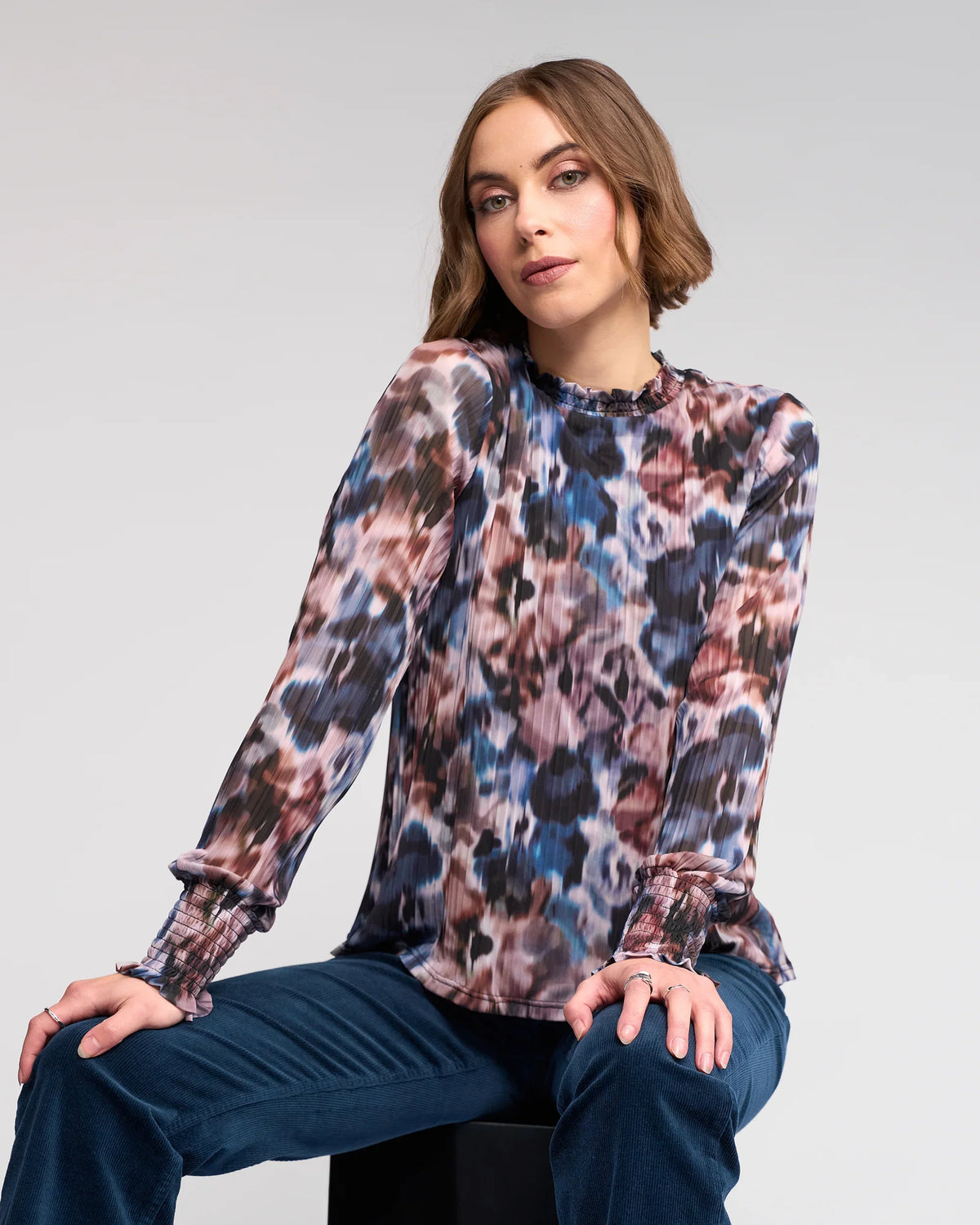 A woman with short brown hair wears the Knewe Label Zimi Top - Celine, featuring a long-sleeve abstract print, and blue pants as she sits confidently on a black stool against a light gray background.