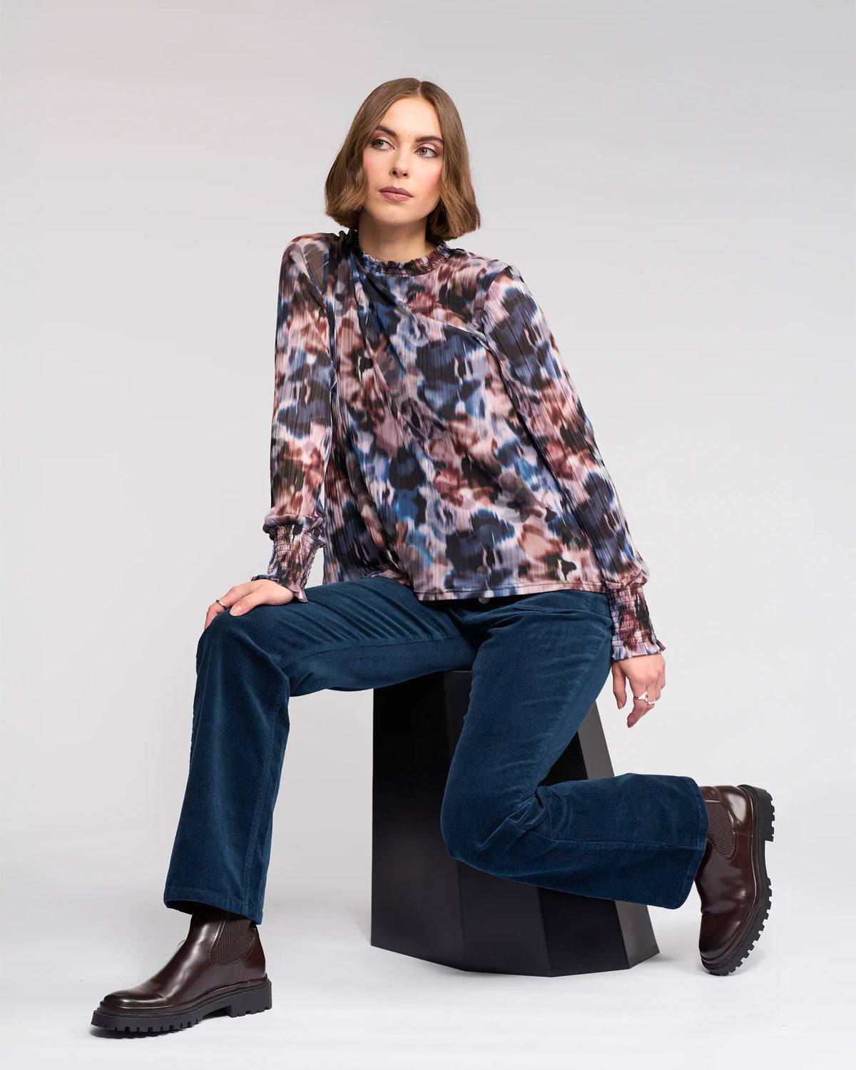 A woman with shoulder-length hair wears the Knewe Label Zimi Top - Celine, featuring multicolored abstract prints, paired with blue corduroy pants and brown boots. She sits on a black geometric stool, looking to the side against a plain background.