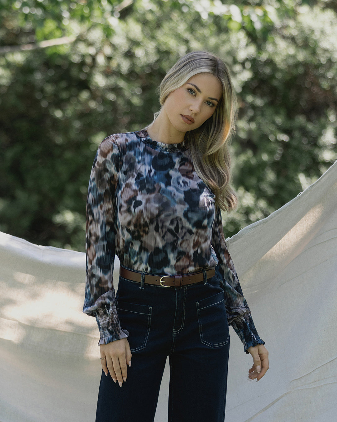 A woman with long, wavy blonde hair stands outdoors in front of a cream-colored sheet. She wears a patterned, long-sleeve blouse and high-waisted dark jeans, with greenery blurred in the background.