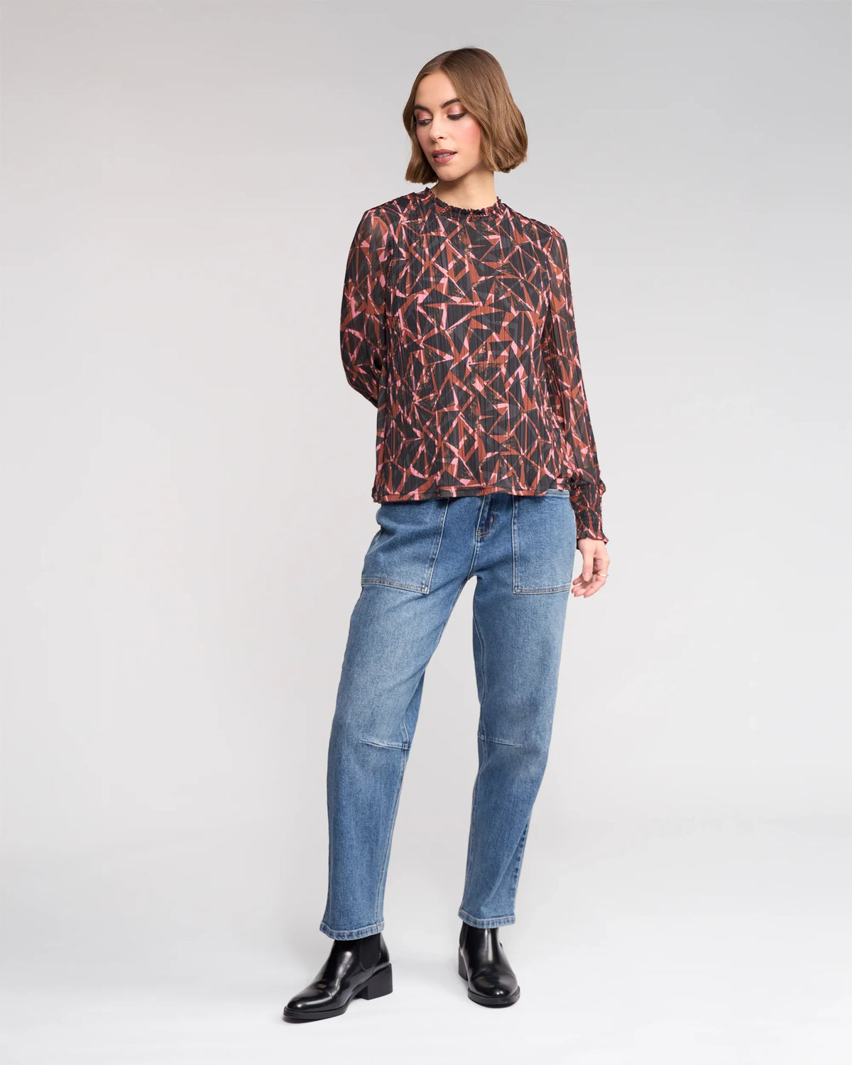A woman with short brown hair stands against a plain background wearing the Knewe Label Zimi Top - Dusk, relaxed blue jeans, and black ankle boots. She looks slightly to the side with one hand behind her back.
