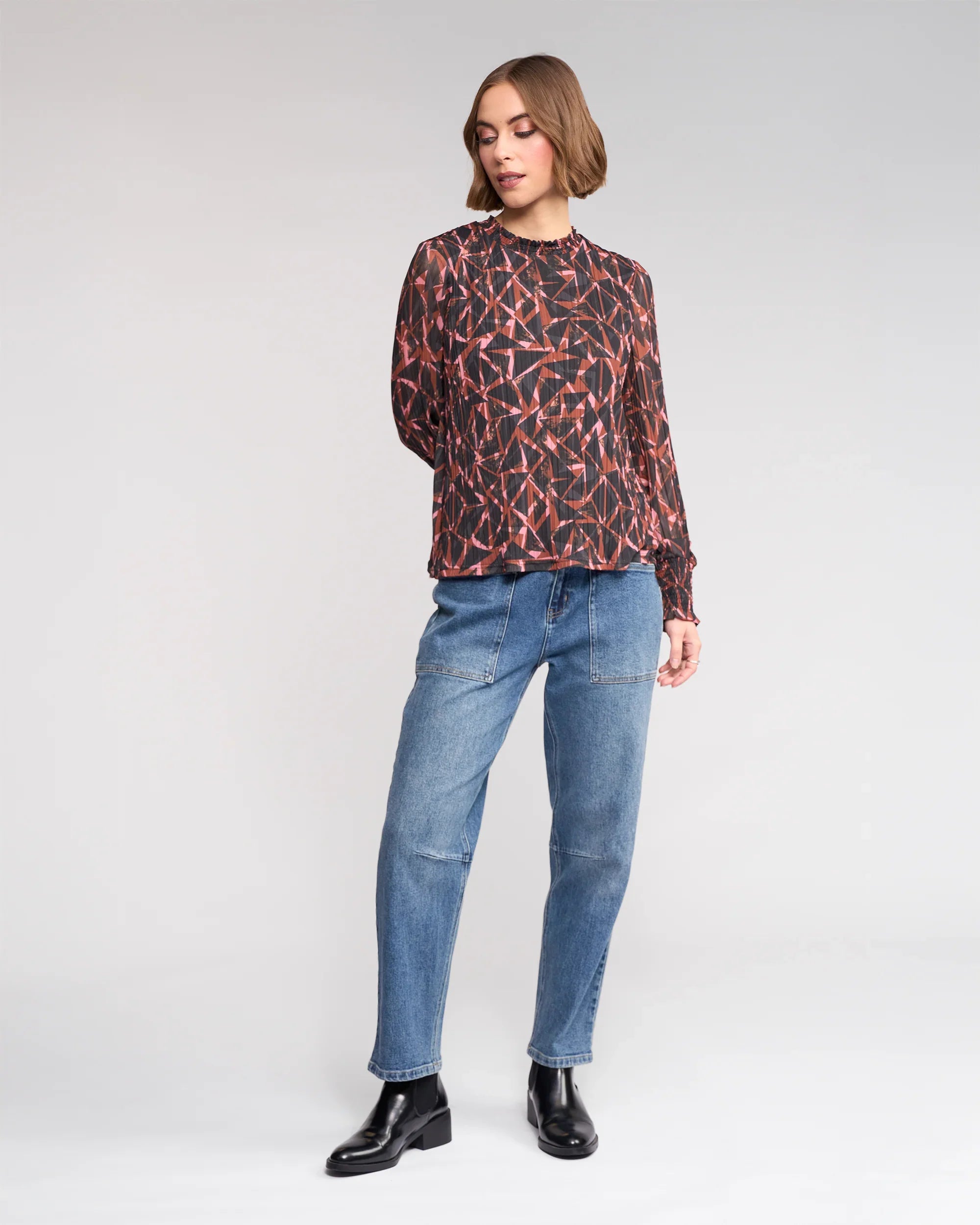 A woman with short brown hair stands against a plain background wearing the Knewe Label Zimi Top - Dusk, relaxed blue jeans, and black ankle boots. She looks slightly to the side with one hand behind her back.