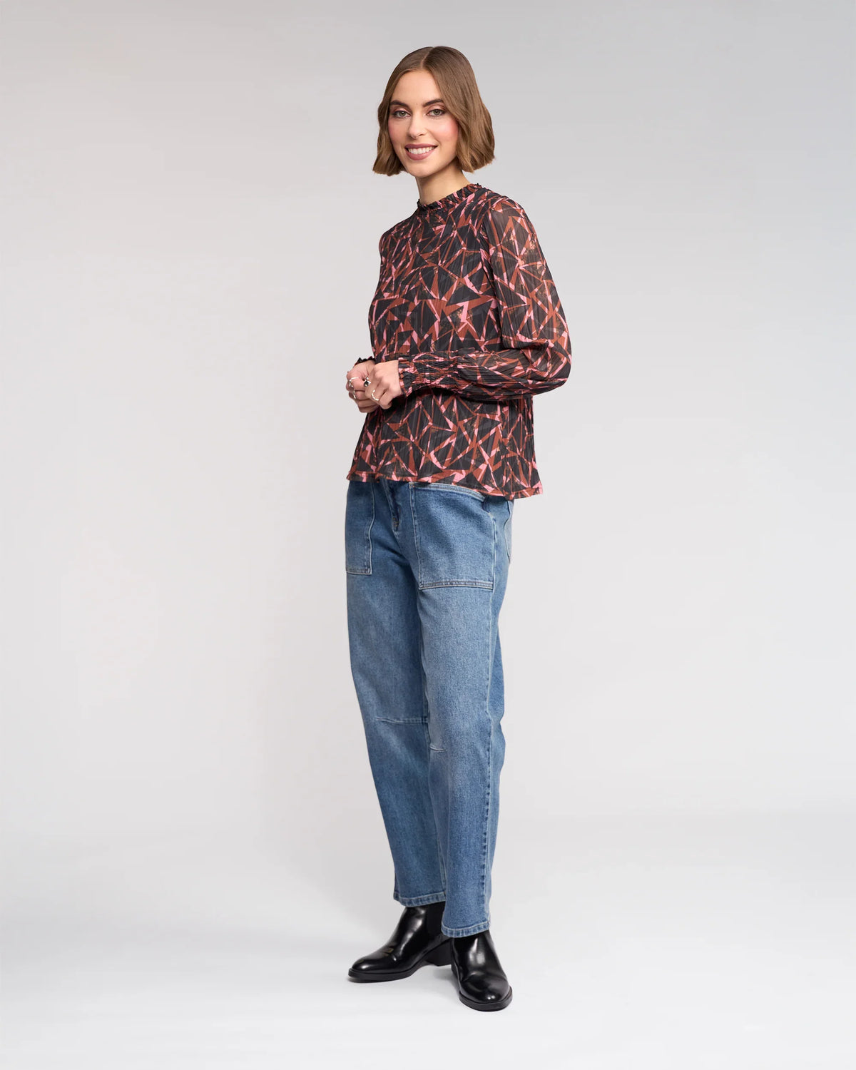 A woman with short brown hair smiles, wearing the Knewe Label Zimi Top - Dusk, blue jeans, and black ankle boots against a plain light gray background.