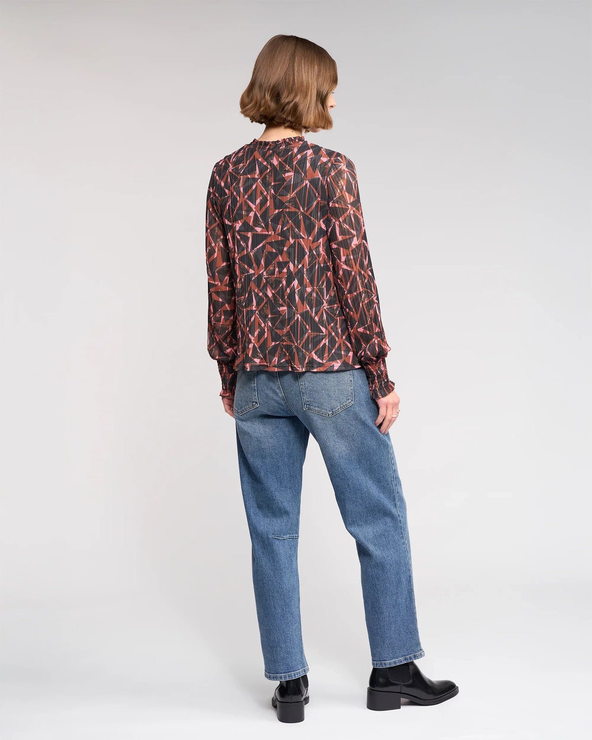 A person with chin-length brown hair, dressed in the Knewe Label Zimi Top - Dusk, blue jeans, and black boots, stands facing away against a plain light background.
