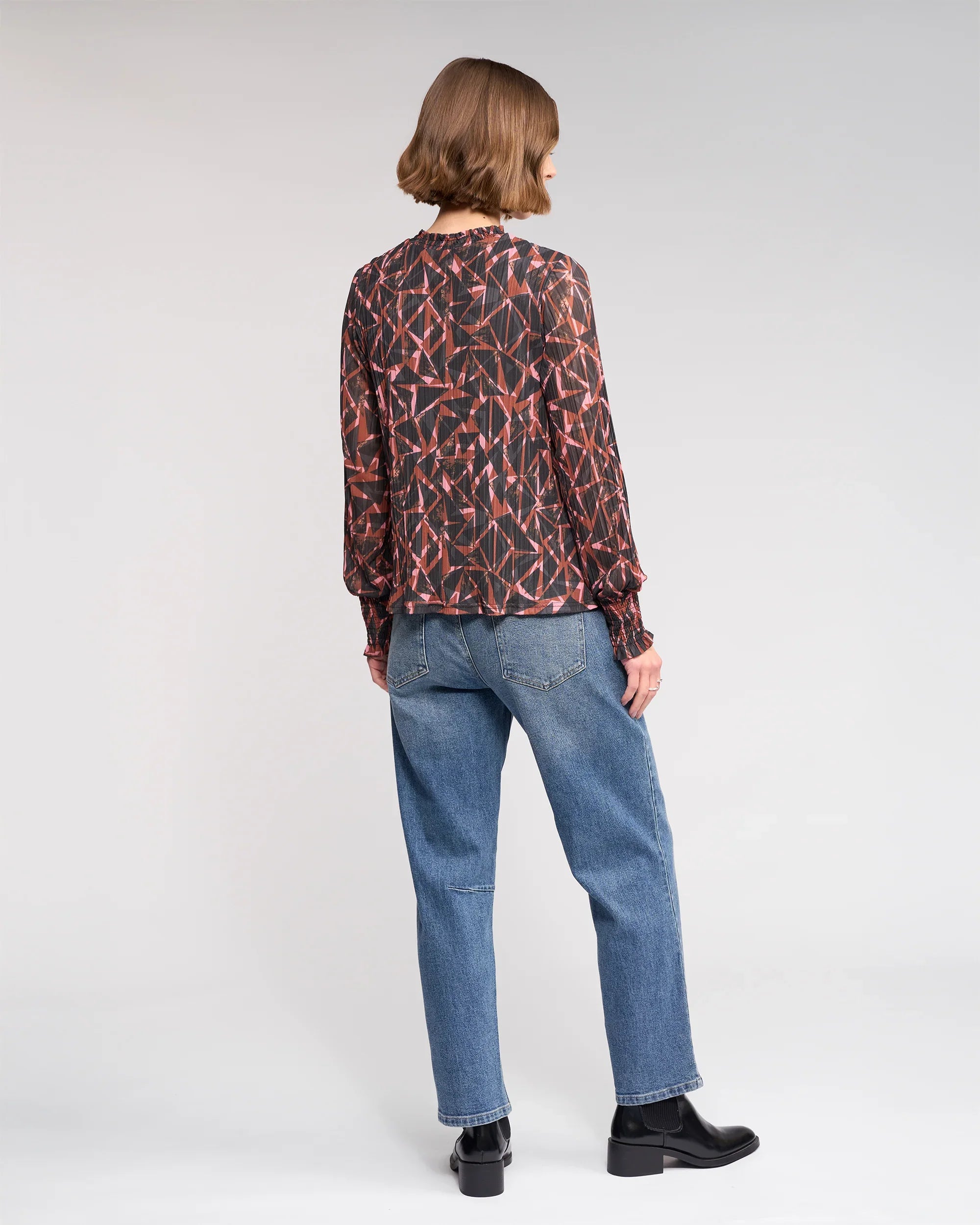 A person with chin-length brown hair, dressed in the Knewe Label Zimi Top - Dusk, blue jeans, and black boots, stands facing away against a plain light background.