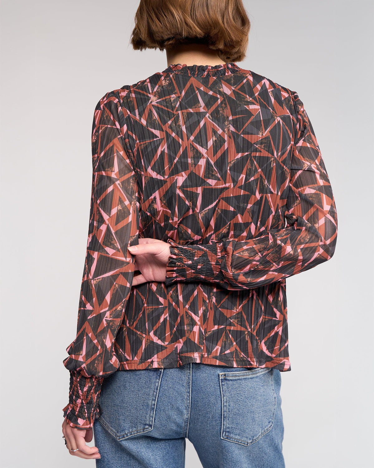 A person with short brown hair is shown from the back, wearing the Knewe Label Zimi Top - Dusk, a long sleeve mesh top with a geometric red and black print, paired with blue jeans.