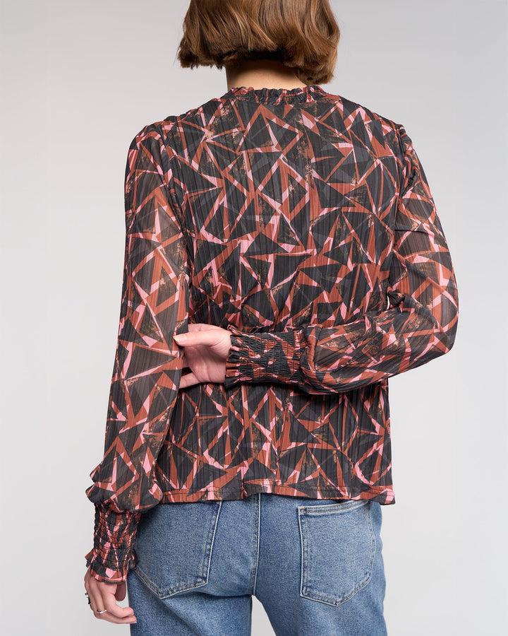 A person with short brown hair is shown from the back, wearing the Knewe Label Zimi Top - Dusk, a long sleeve mesh top with a geometric red and black print, paired with blue jeans.