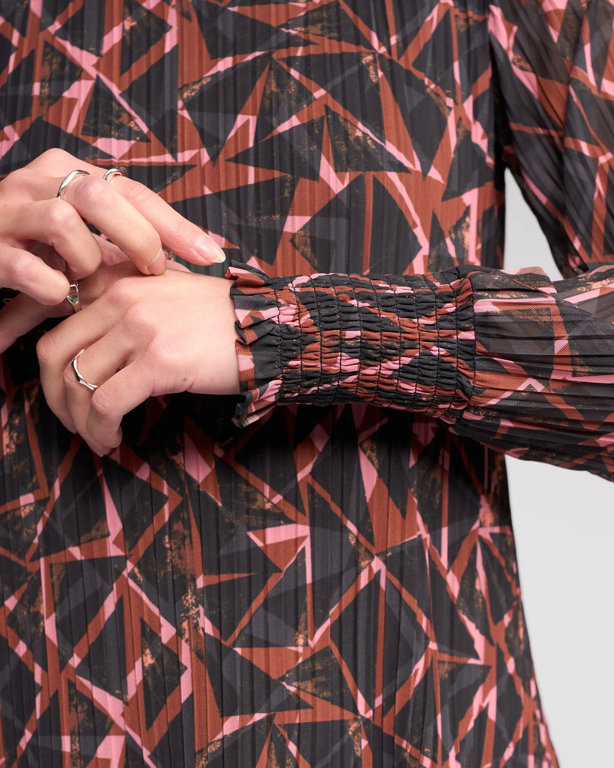 A close-up of someone adjusting the cuff of the Knewe Label Zimi Top – Dusk, a long sleeve top featuring a brown, black, and pink print. Multiple thin rings adorn their fingers.