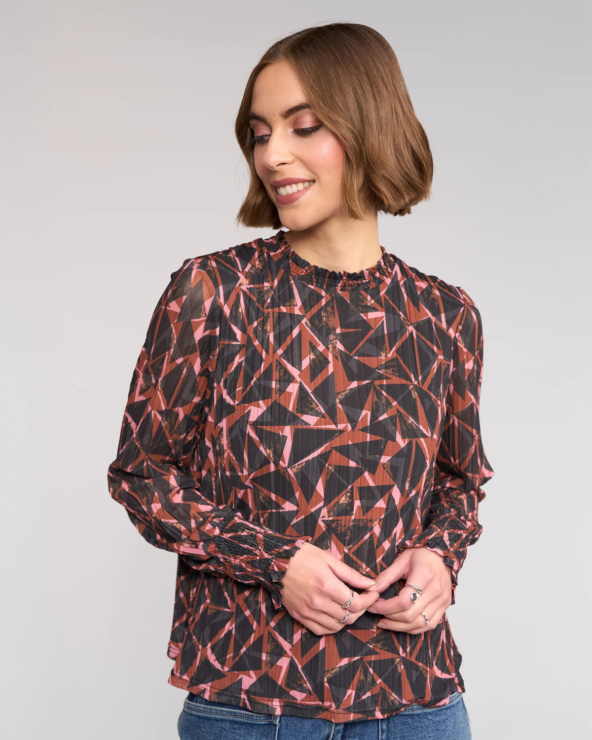 A woman with short brown hair wears the Knewe Label Zimi Top – Dusk, featuring bold red and black geometric shapes, as she stands against a plain light background, smiling and looking down to her left.