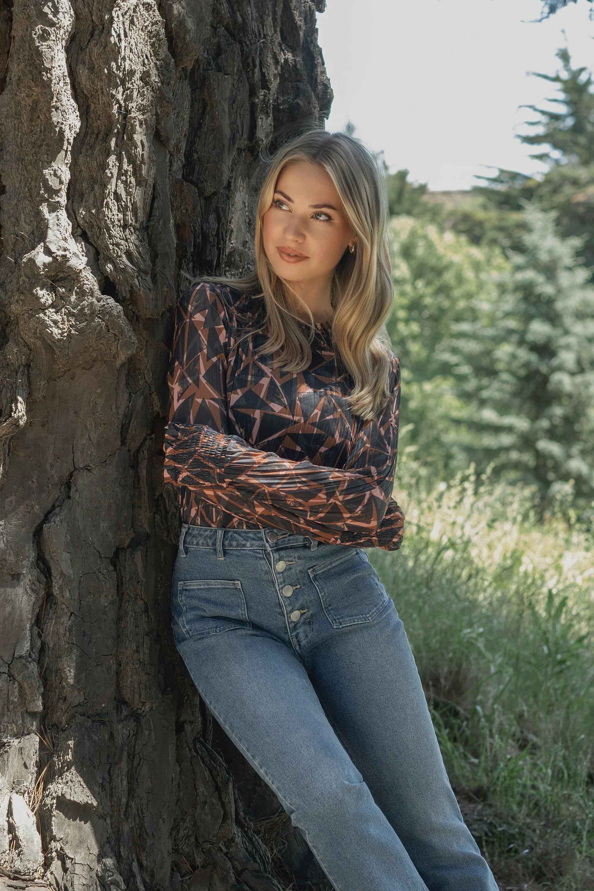 A woman with long blonde hair leans against a tree outdoors, wearing the Knewe Label Zimi Top in Dusk and high-waisted blue jeans, surrounded by greenery and trees.