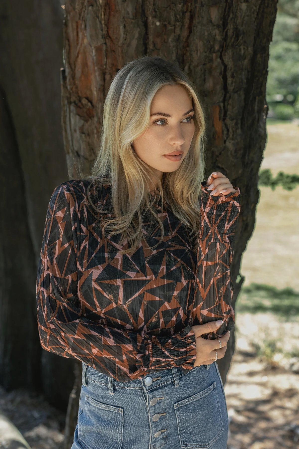 A woman with long blonde hair stands by a tree wearing the Knewe Label Zimi Top – Dusk and high-waisted blue jeans, gazing thoughtfully to the side.