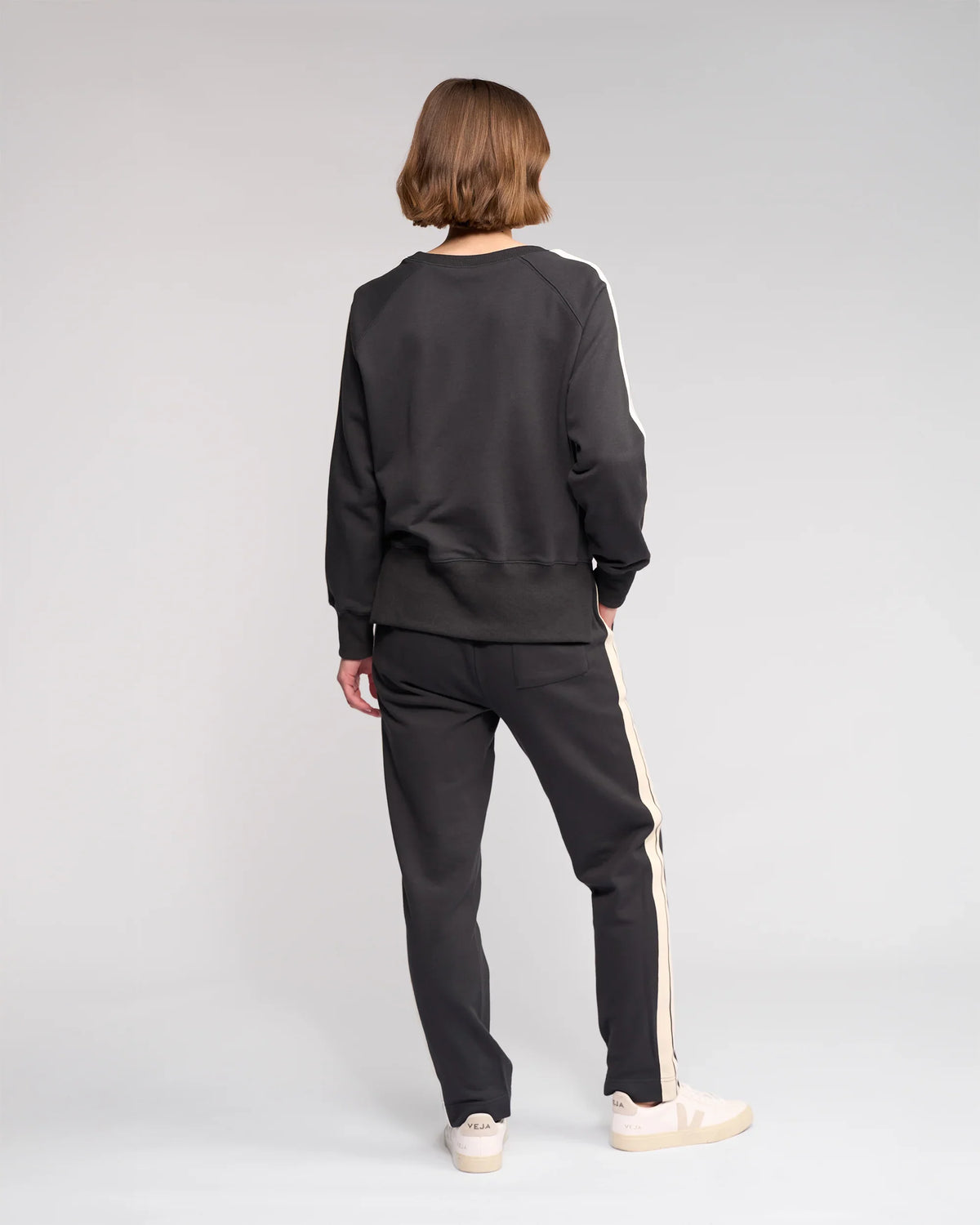 A person with short hair stands facing away, wearing a black sweatshirt and black pants with white stripes down the sides, and white sneakers, against a plain light gray background.