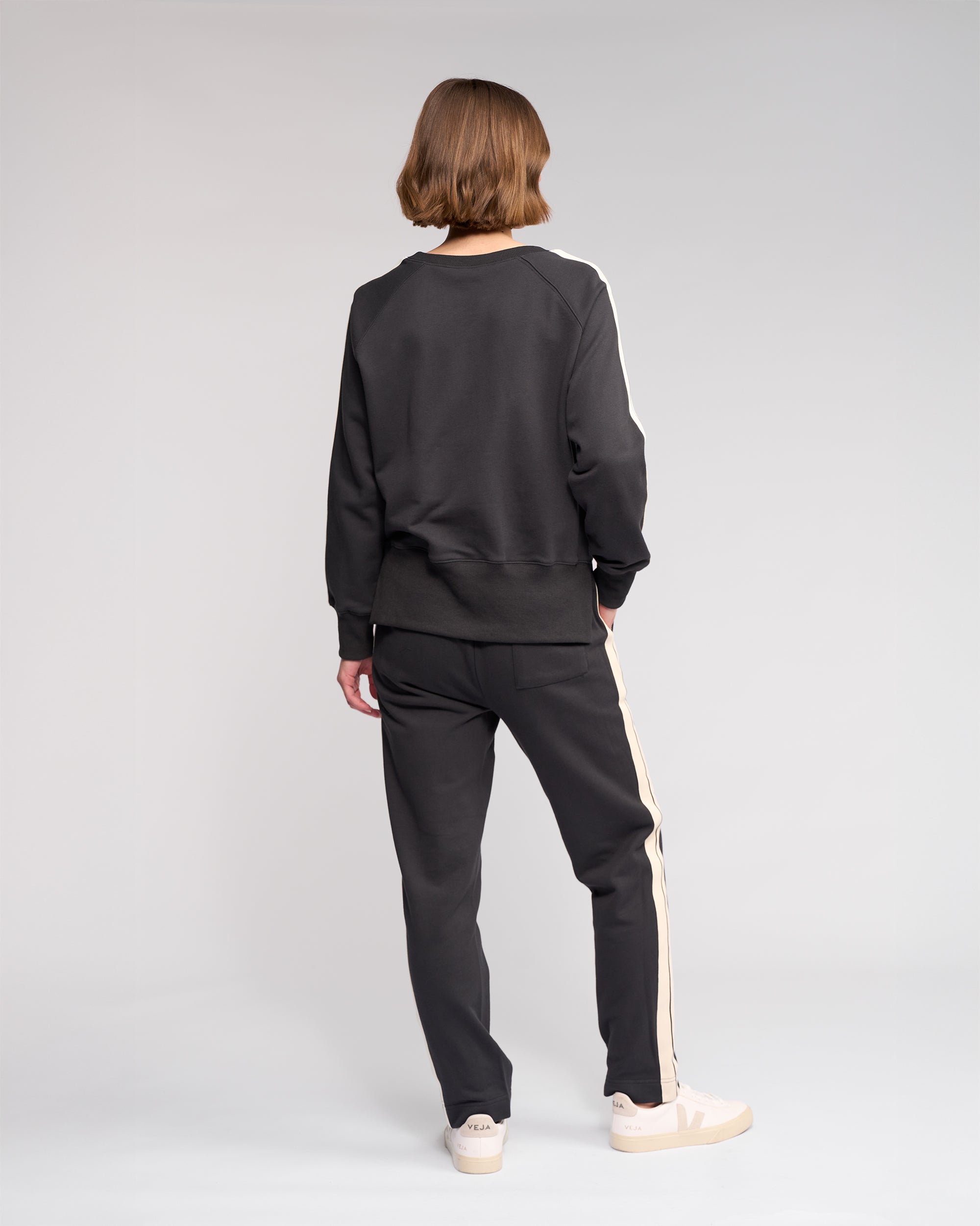 A person with short hair stands facing away, wearing a black sweatshirt and black pants with white stripes down the sides, and white sneakers, against a plain light gray background.