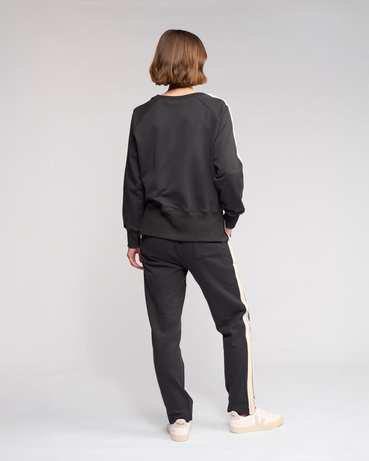 A person with short hair stands facing away, wearing a black sweatshirt and black pants with white stripes down the sides, and white sneakers, against a plain light gray background.