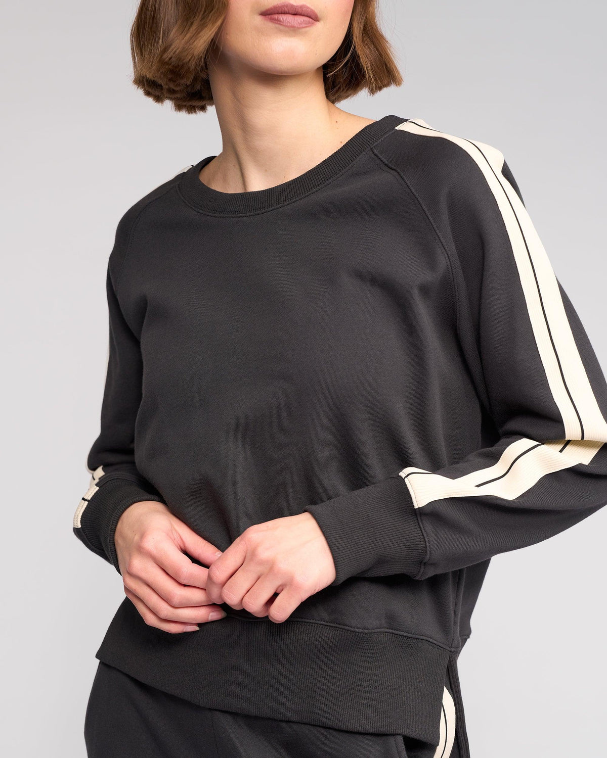 A woman wearing a black sweatshirt with white stripes on the sleeves stands against a plain background, shown from the shoulders to the hips. Her hands are clasped near her waist.