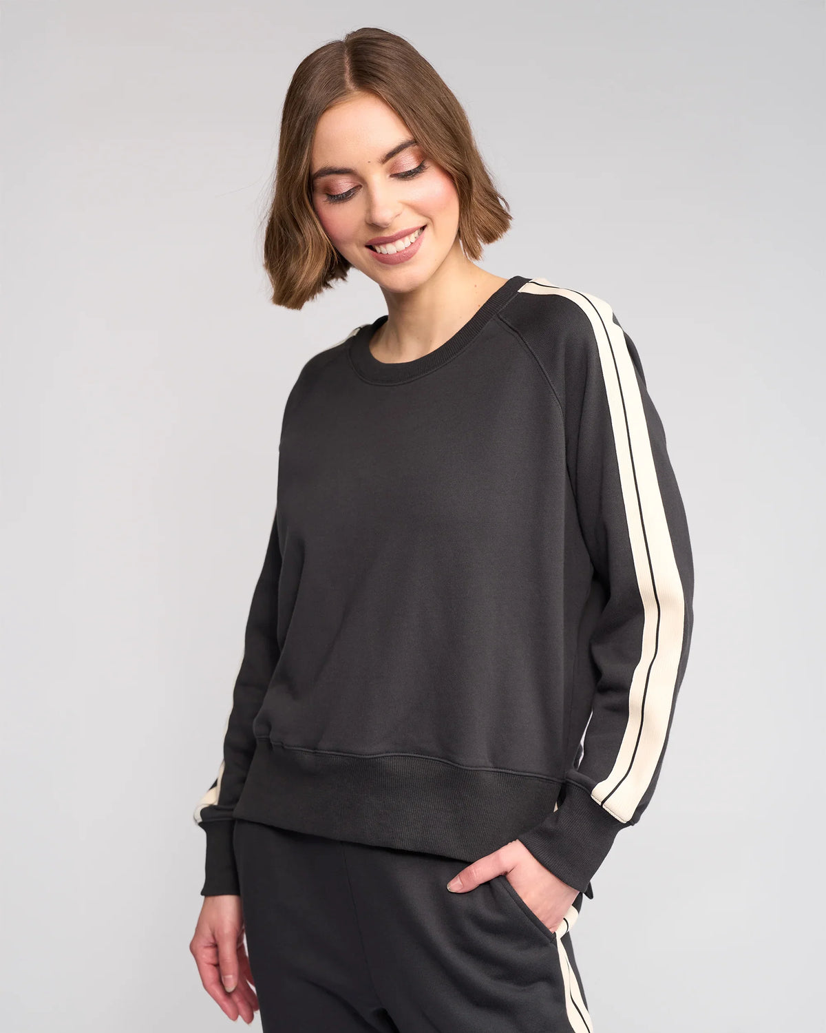 A woman with short brown hair is smiling and looking down while wearing a black sweatshirt with white stripes on the sleeves and matching black pants. The background is plain and light-colored.