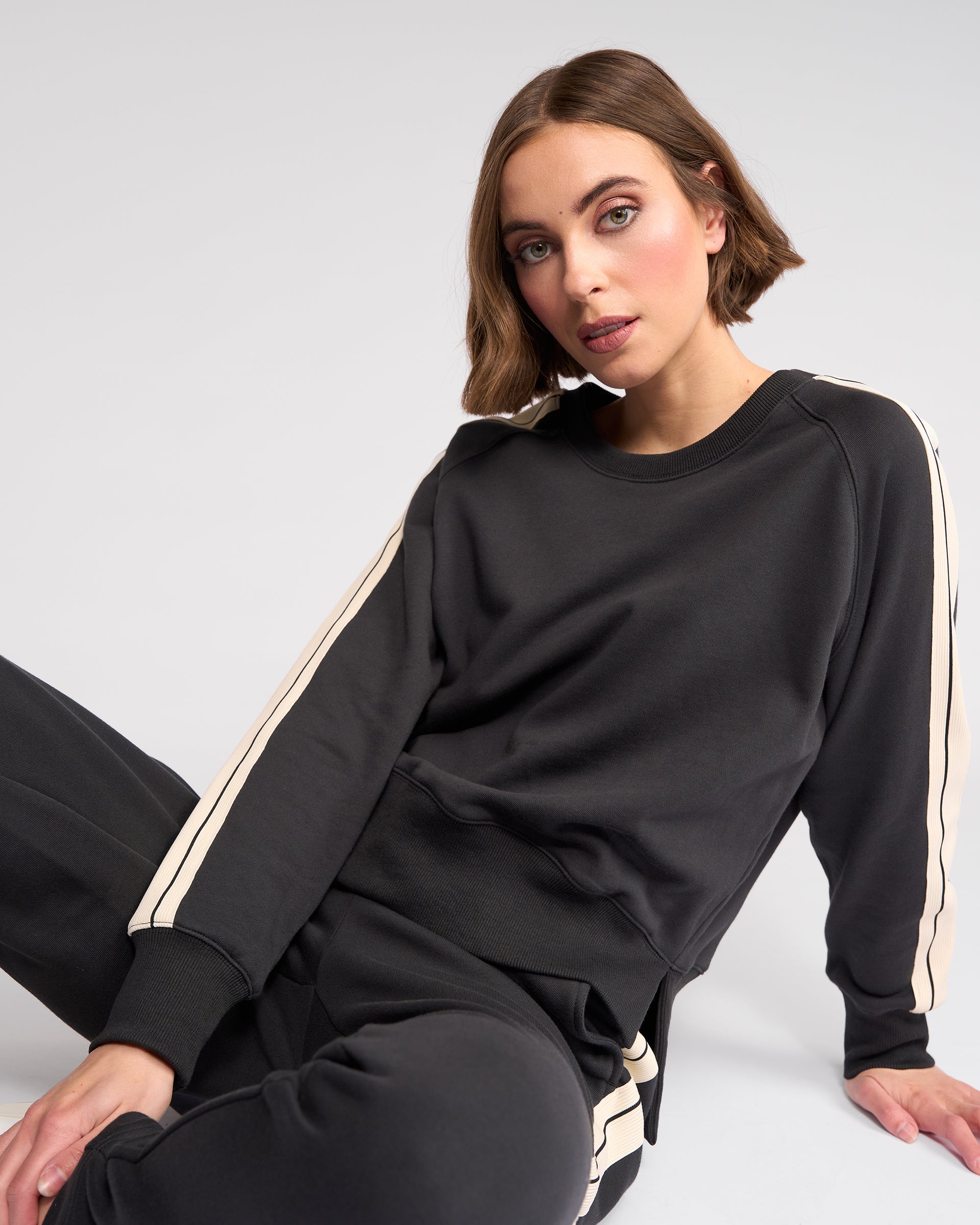 A woman with short brown hair, wearing a black sweatshirt and matching pants with white stripes down the sleeves and legs, is sitting on the floor against a plain light background, looking toward the camera.