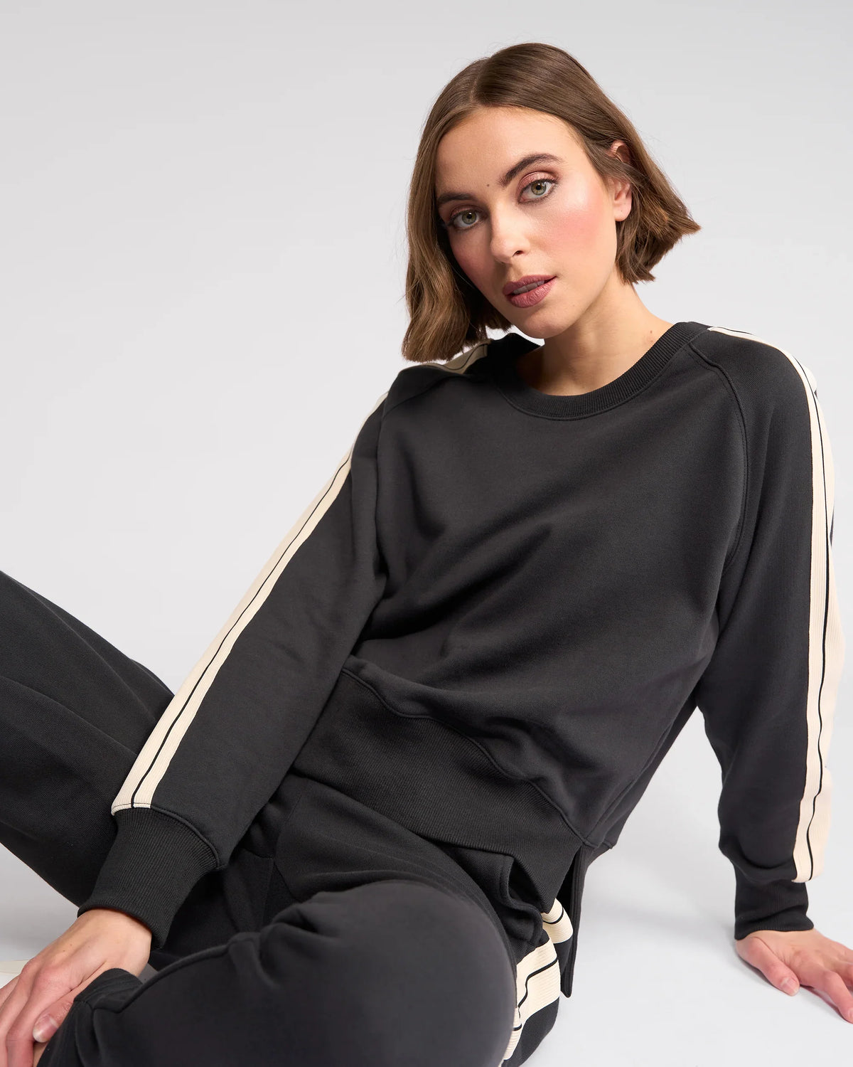 A woman with short brown hair, wearing a black sweatshirt and matching pants with white stripes down the sleeves and legs, is sitting on the floor against a plain light background, looking toward the camera.